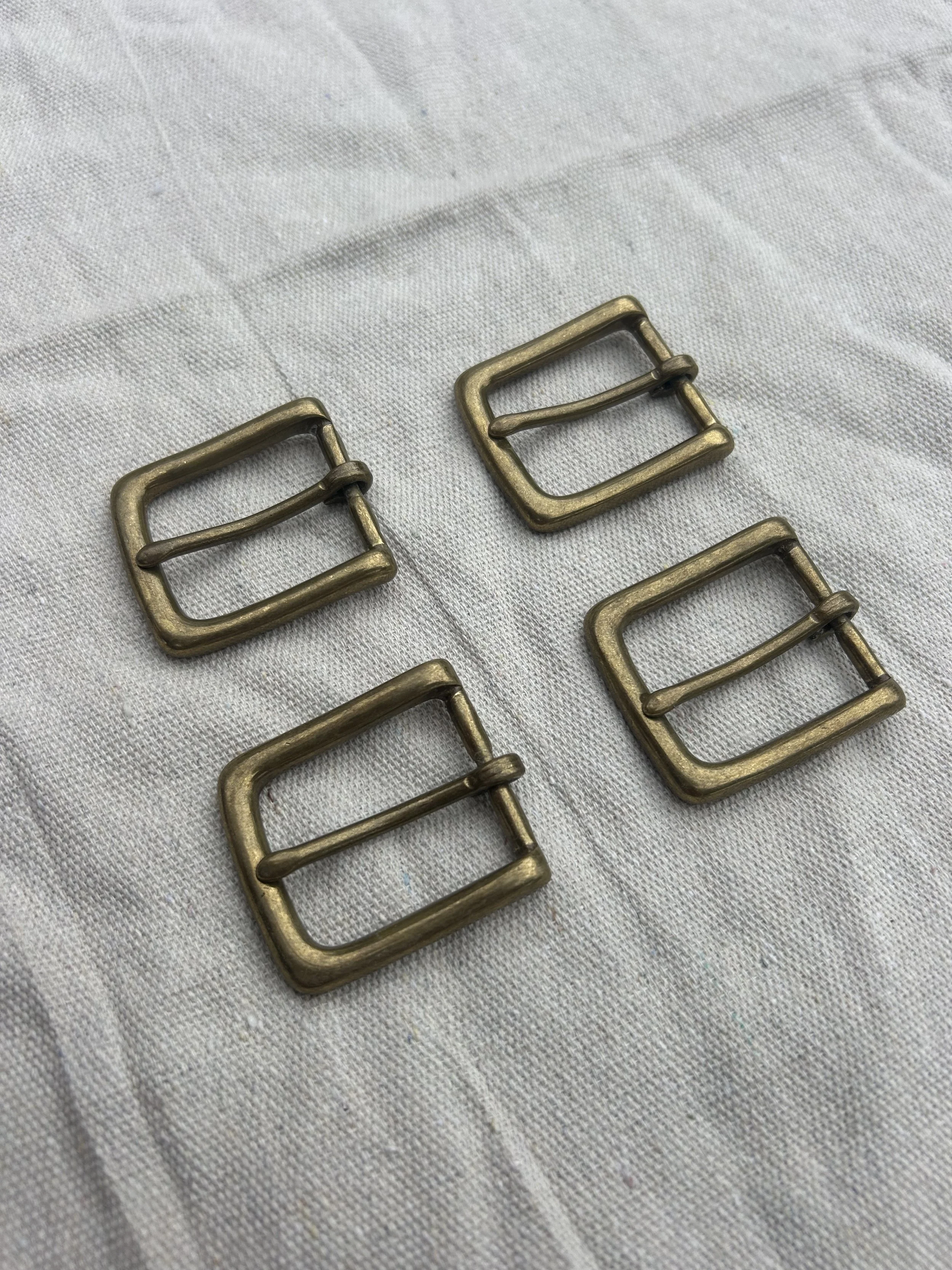 Four brass rectangular buckles with a central bar, placed on a white fabric background.