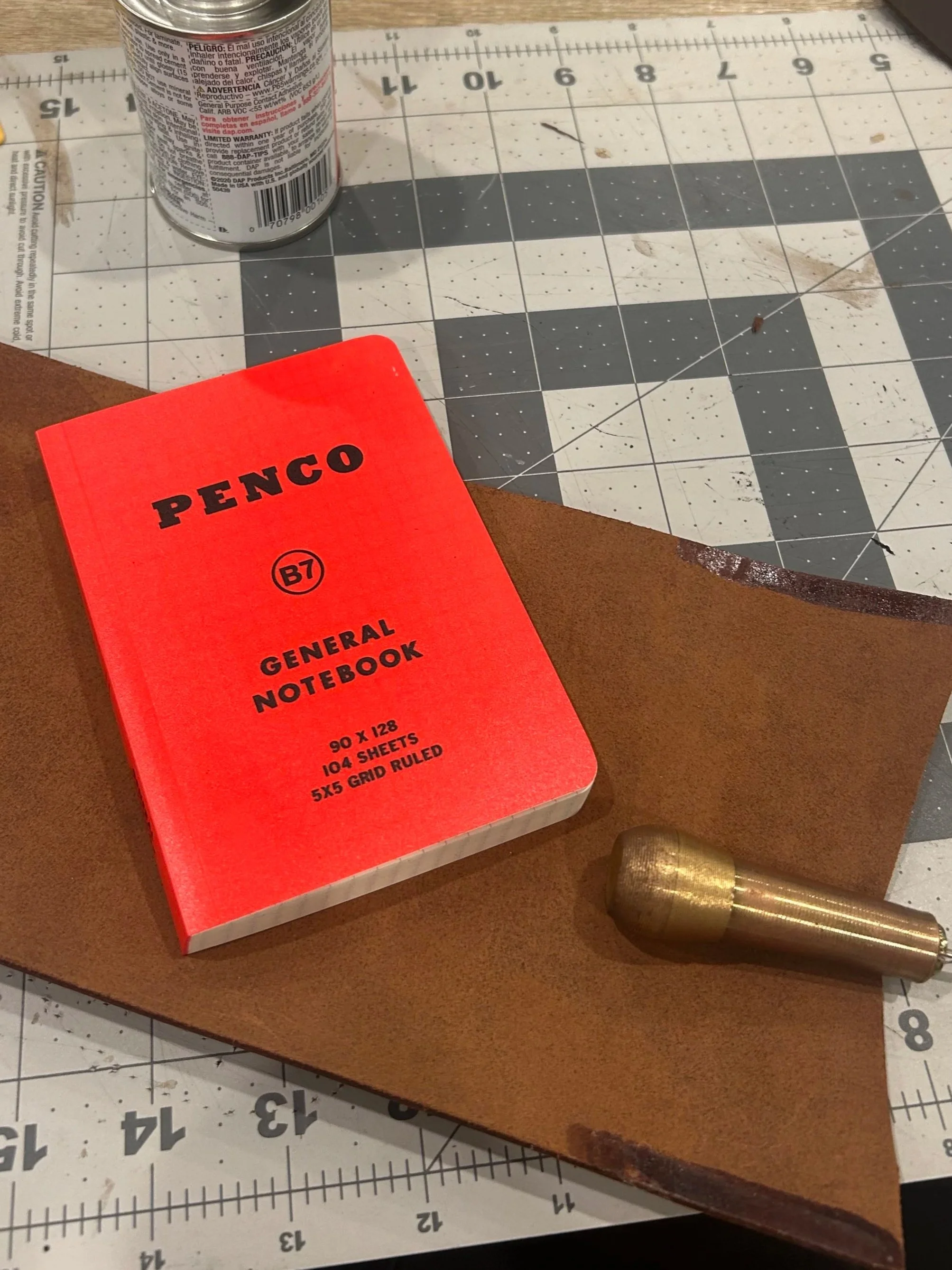 A red Penco general notebook, a brass hammer, a piece of brown leather, and a can of spray paint on a cutting mat with measurement markings.