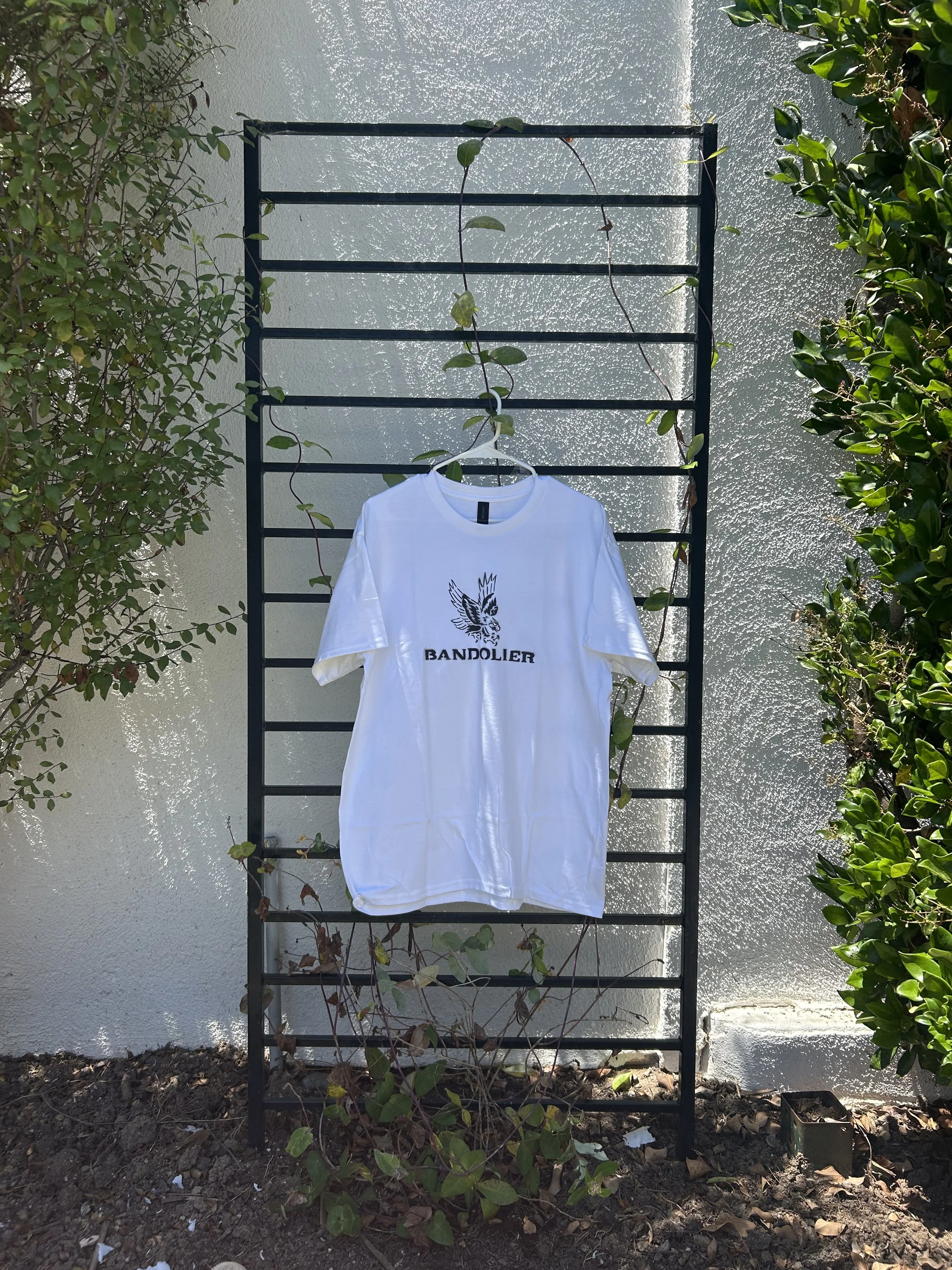 White T-shirt with a black eagle graphic and the text 'BANDOHLER' hanging on a hanger on a metal outdoor garden trellis with green plants on either side.