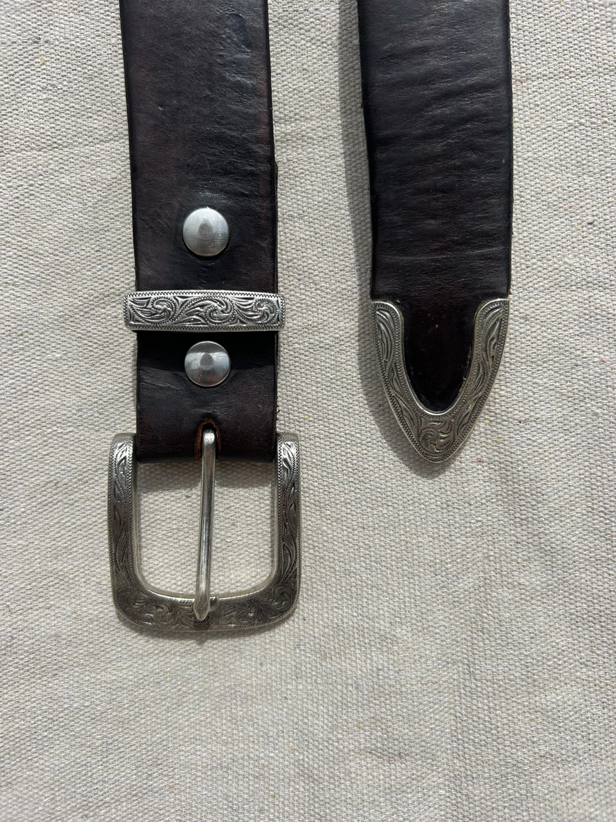 A black leather belt with silver engraved decorative hardware, including a rectangular buckle, a belt loop, and a tip with intricate patterns.