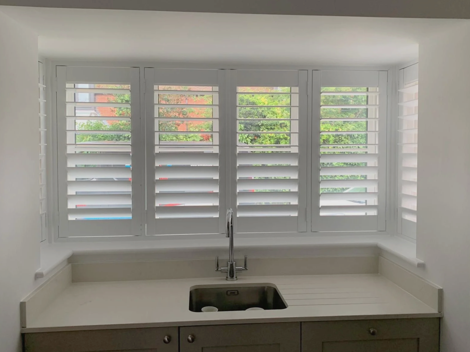 Kitchen window with white plantation shutters above a white countertop and a stainless steel sink.