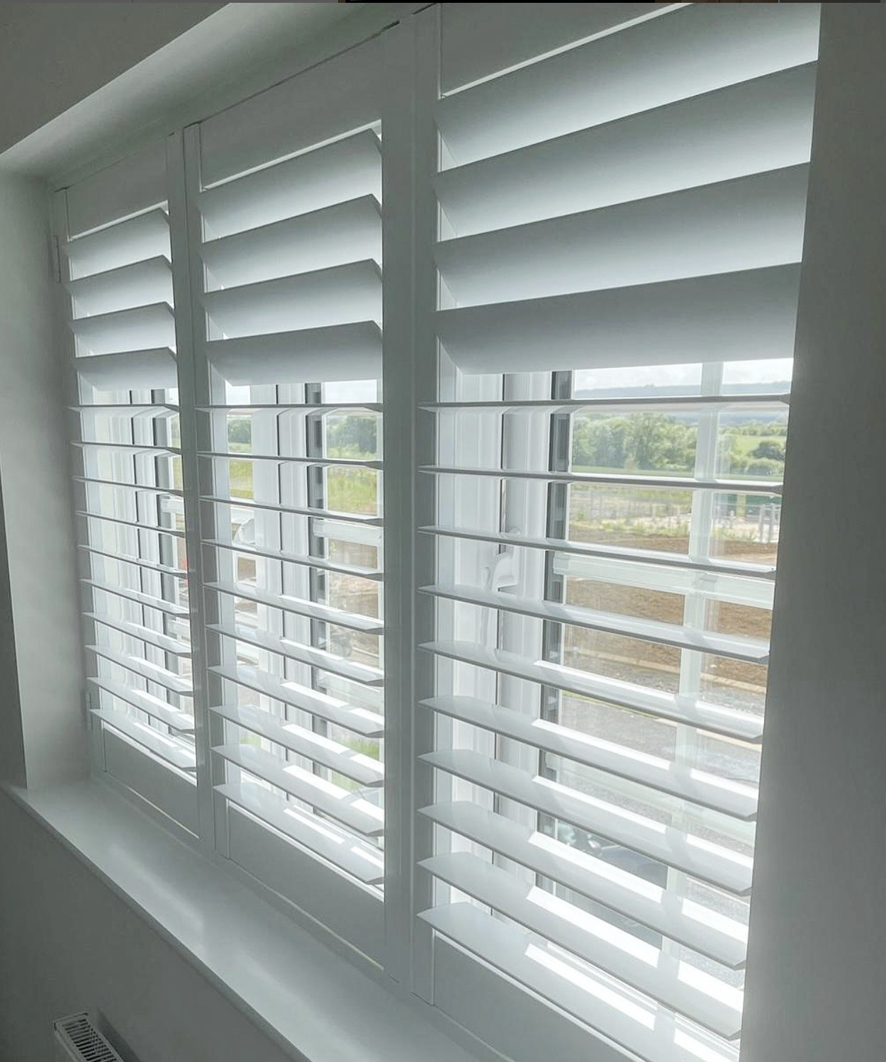 White plantation shutters on a window, partially open, showing a view of trees and a cloudy sky outside.