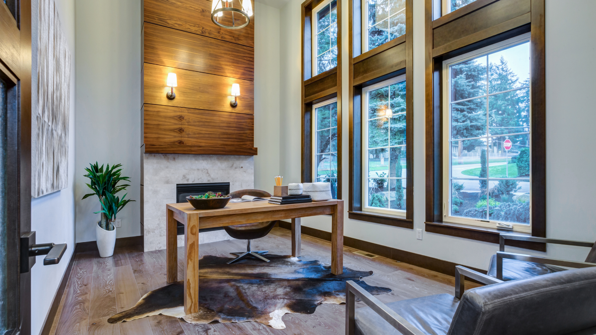 Modern office with large windows, wooden wall paneling, a fireplace, and a wooden desk with a chair, situated on a cowhide rug.