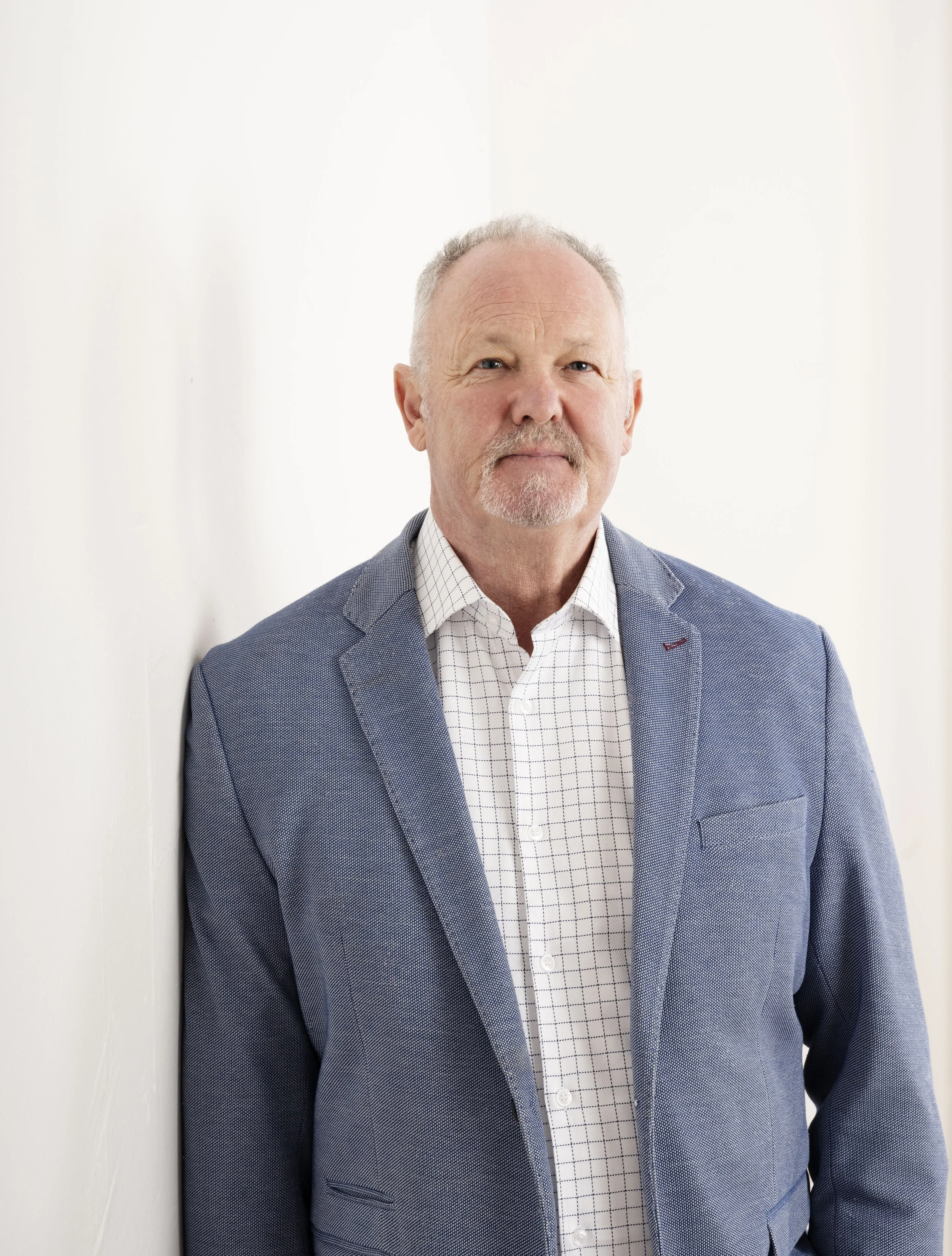 A middle-aged man with short gray hair, a beard, and a mustache, wearing a blue blazer and a white checkered shirt, standing against a plain white background.