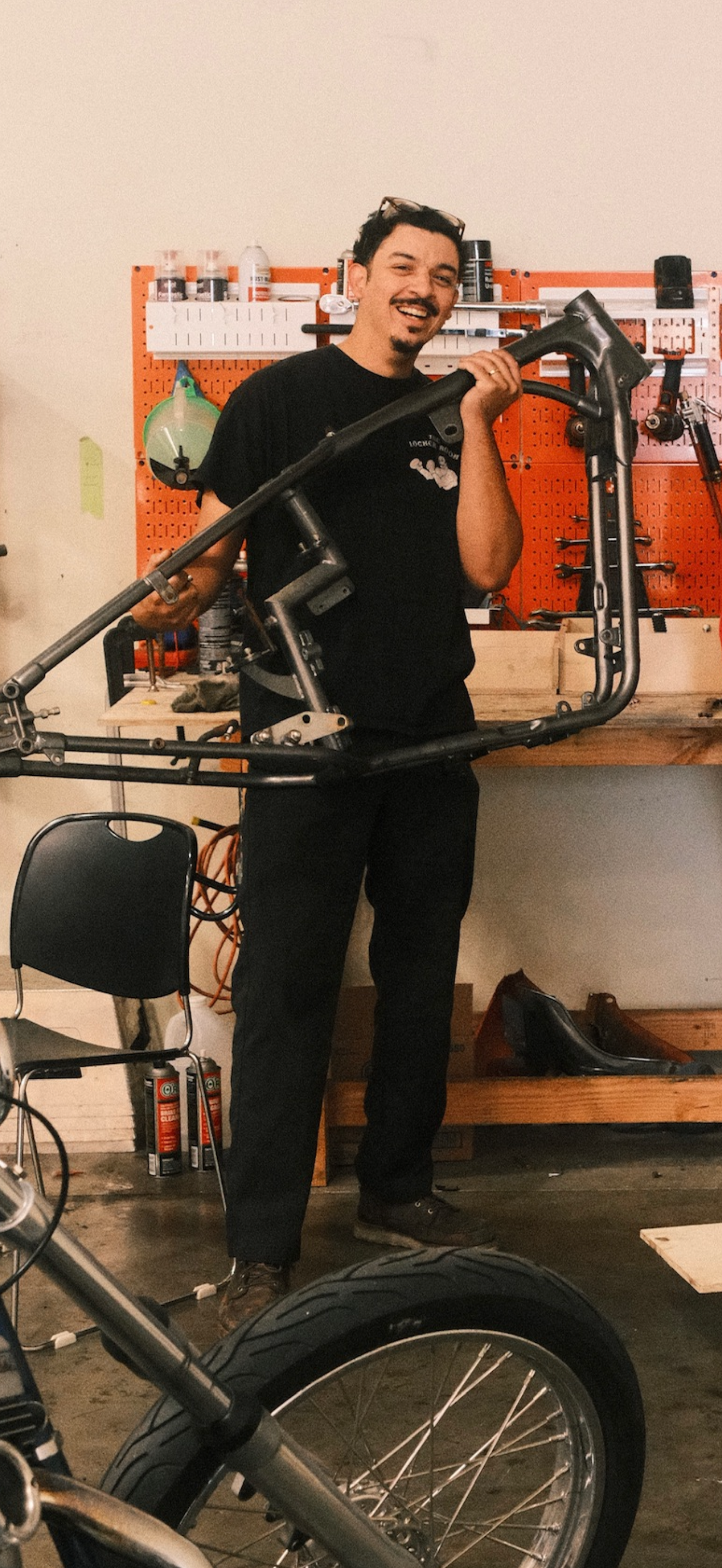 A man in a workshop holding a bicycle frame, smiling.
