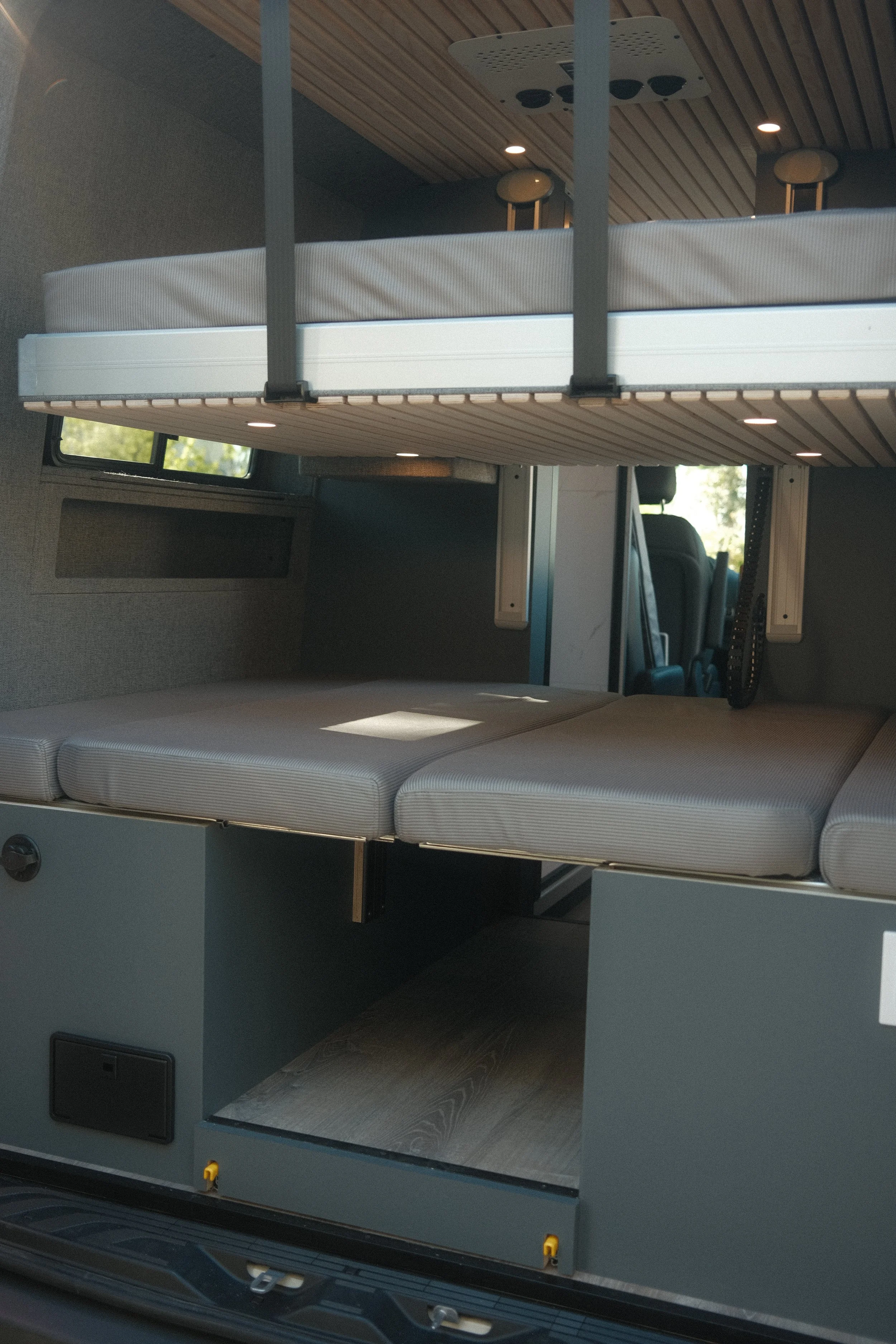 Rear view of a custom interior build for sprinter van featuring modular bench seating that turns into a bed, and a lowered bunk bed above it with garage storage under