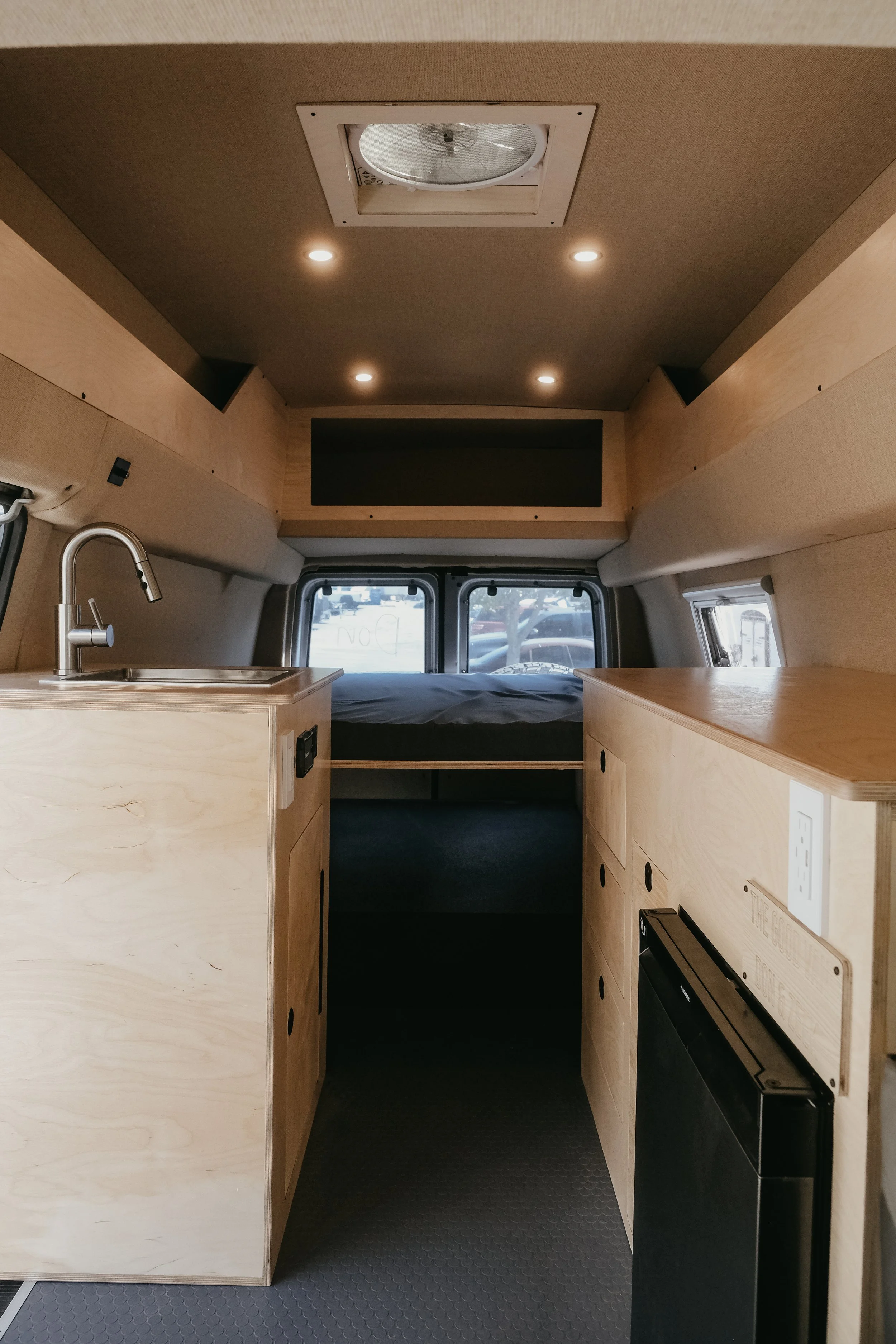 Modern custom build inside sprinter van featuring wood cabinets, sink, mini fridge, bed at the end, and a maxx air vent in the ceiling