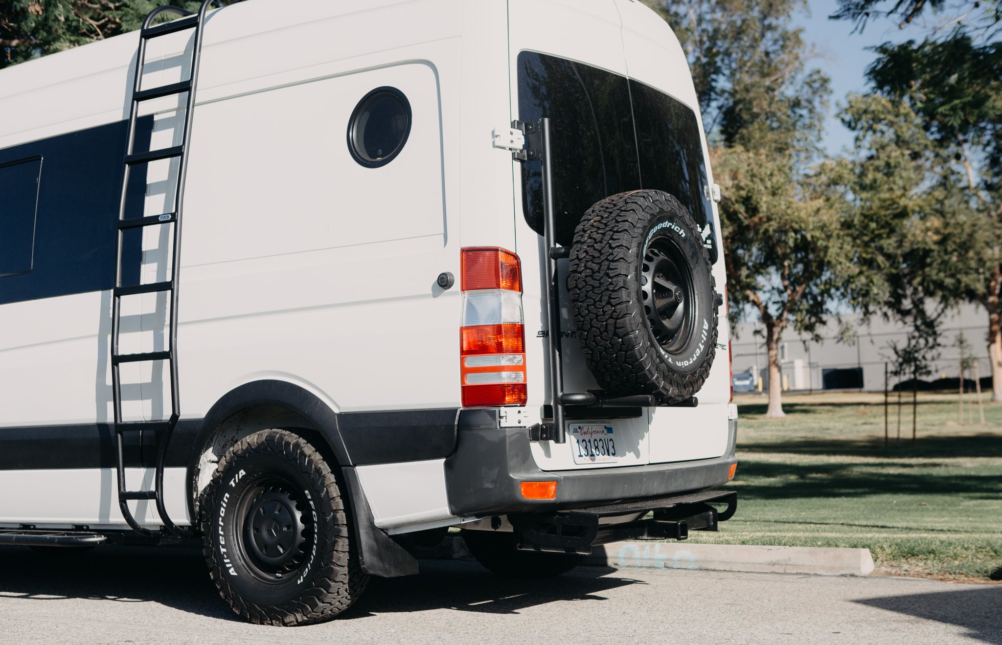 Rear side view of Mercedes sprinter van with side ladder, new tires, lift kit, and tire carrier at the rear