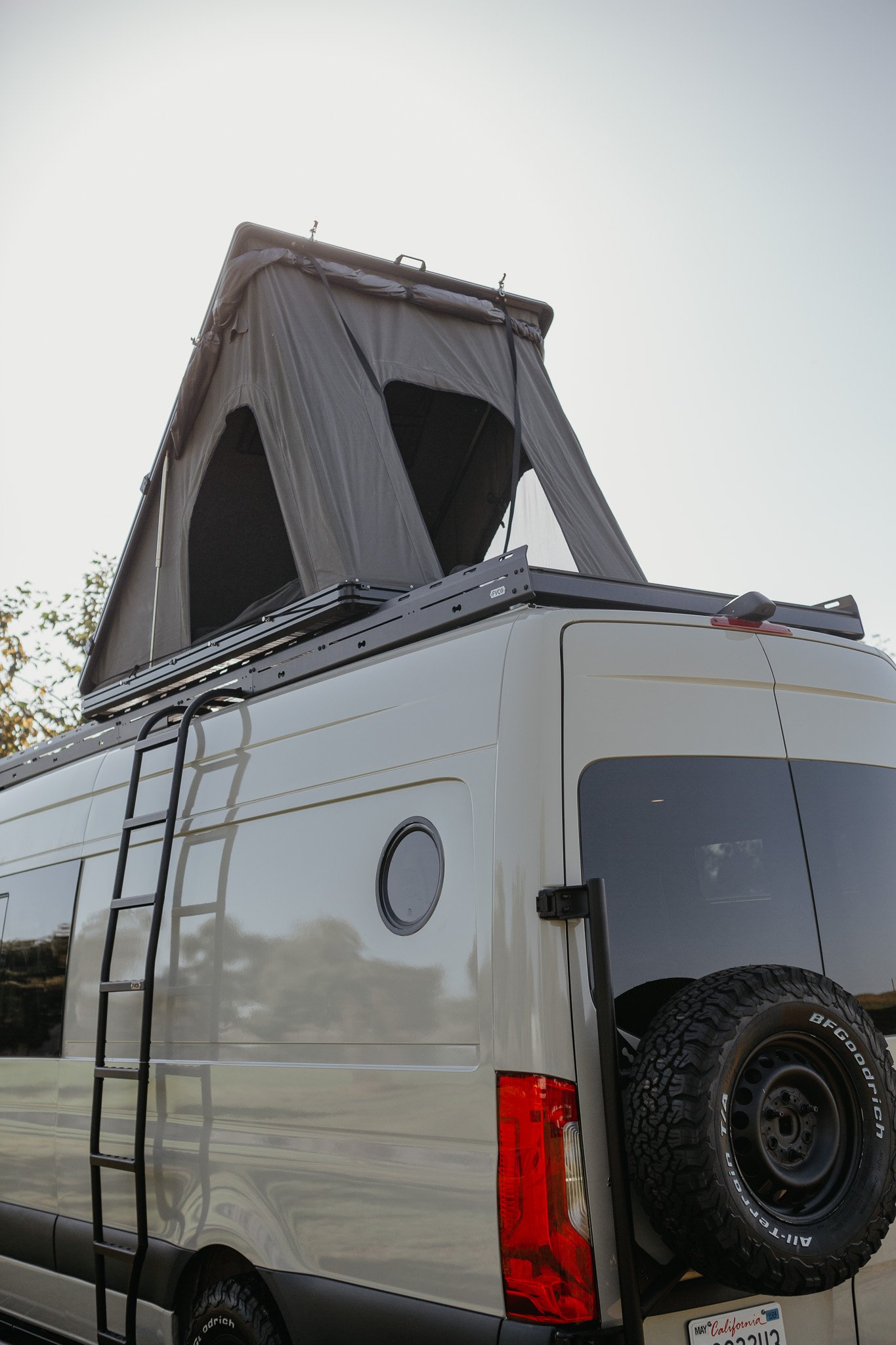 Side view of a mercedes sprinter van with a pop up super pacific tent on the roof with a side ladder, tire carrier, and a custom circular window
