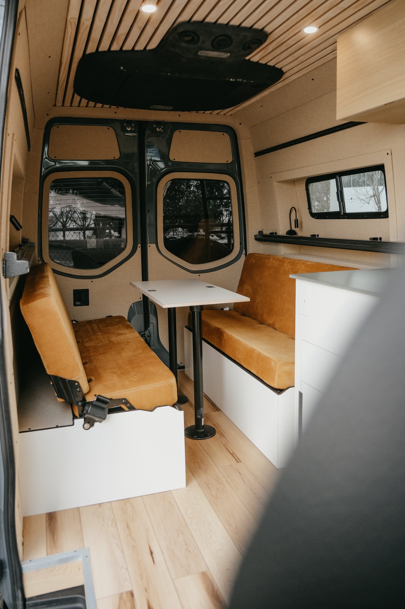 Custom interior build featuring two custom bench seating with a table, ceiling paneling, and wood flooring inside sprinter van. 