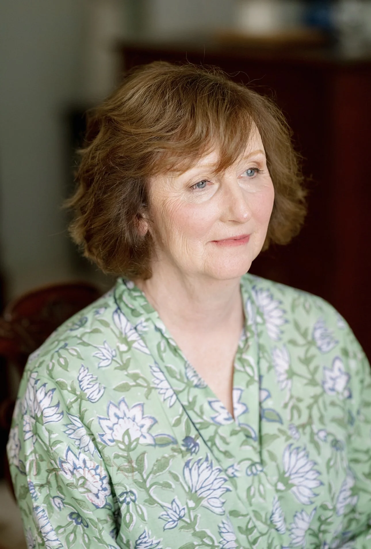Close-up of a middle-aged woman with short brown hair and blue eyes, wearing a green floral blouse, sitting indoors with a blurred background.