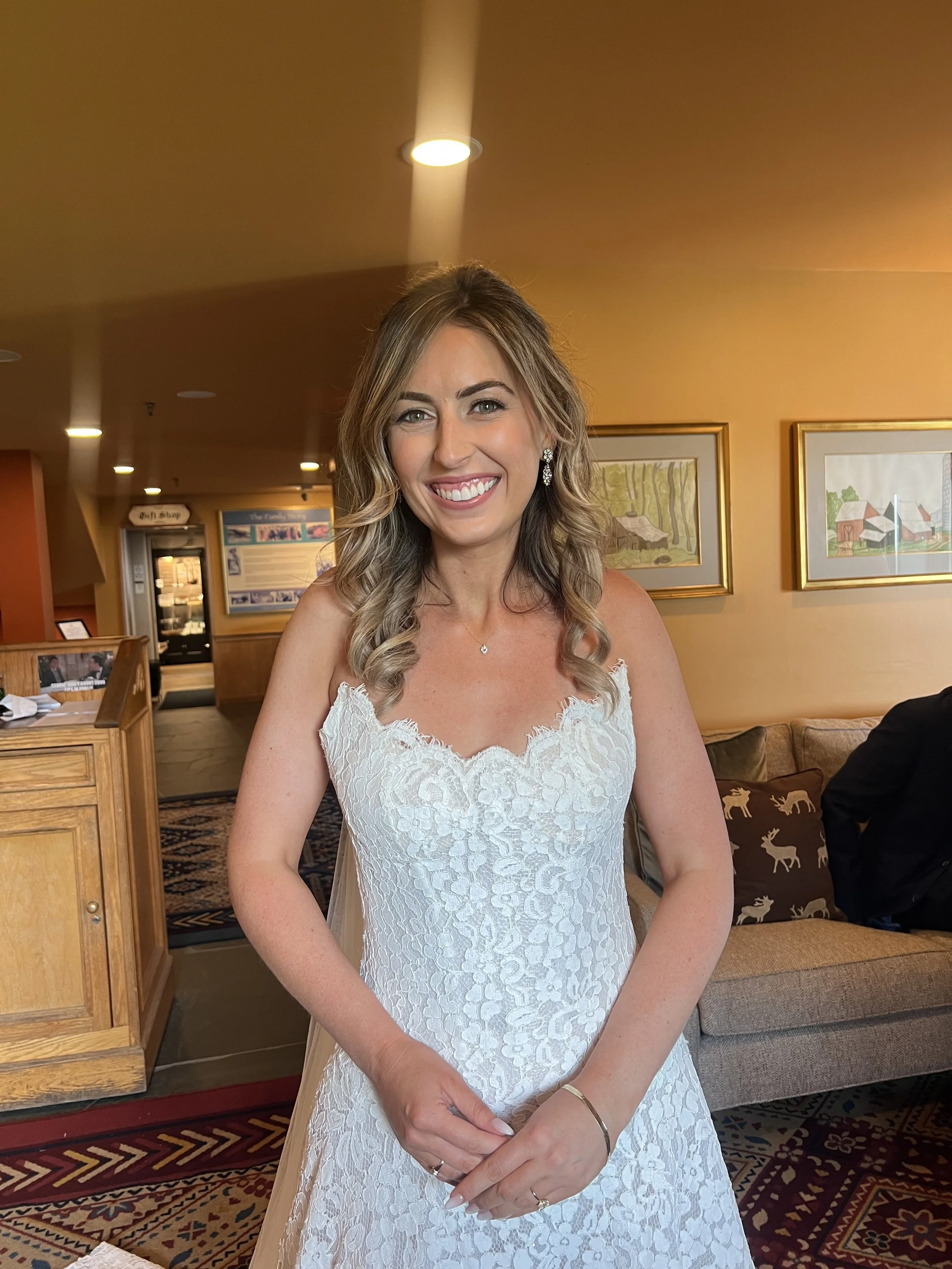 A woman in a white lace dress, smiling at the camera indoors with framed pictures on the wall behind her.
