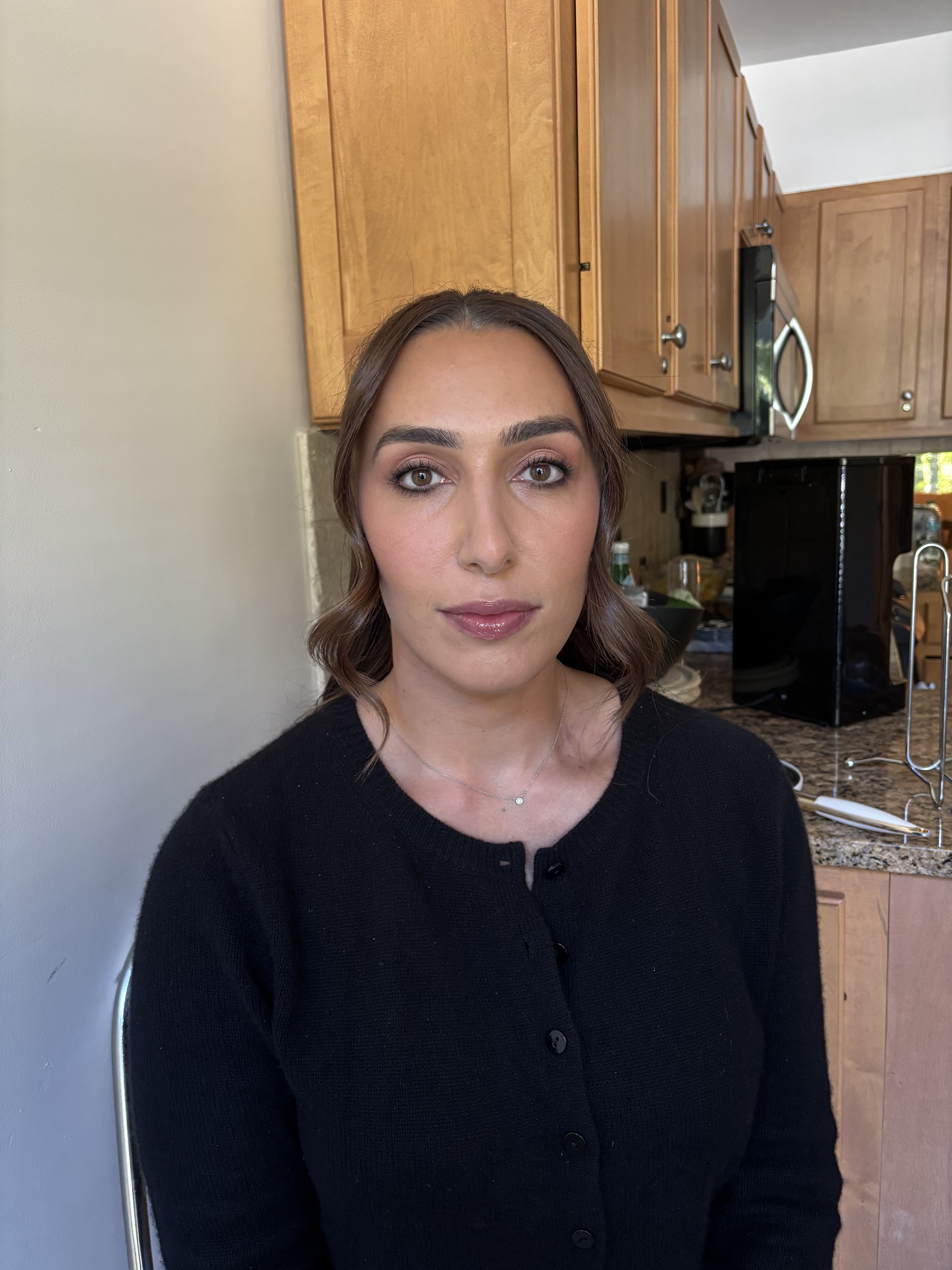 A woman with brown hair and wearing a black button-up sweater sitting in a kitchen with wooden cabinets and granite countertop.