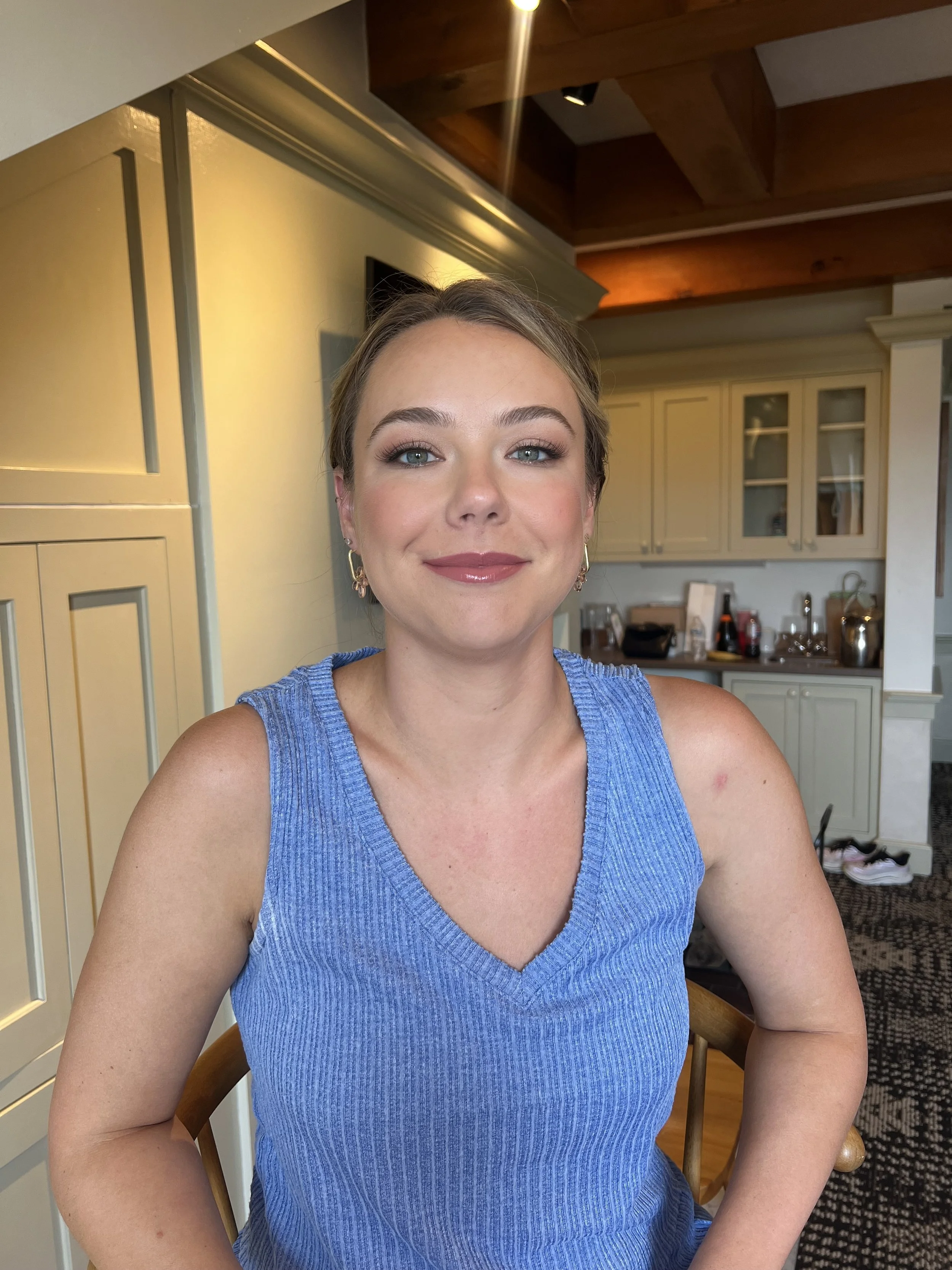 A woman with blonde hair, blue eyes, and makeup, wearing a sleeveless blue top, smiling while sitting in a kitchen with light-colored cabinets and wooden ceiling beams.