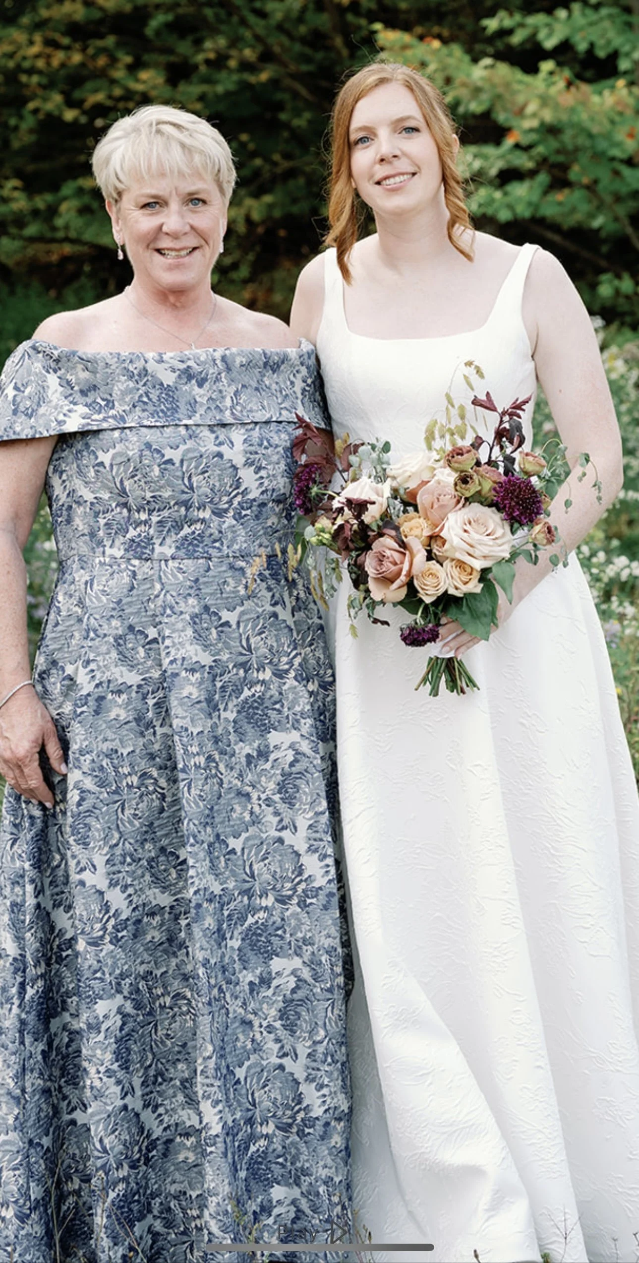 A woman in a white wedding dress holding a bouquet of pink and purple flowers, standing next to an older woman in a blue floral dress, outdoors with green foliage in the background.
