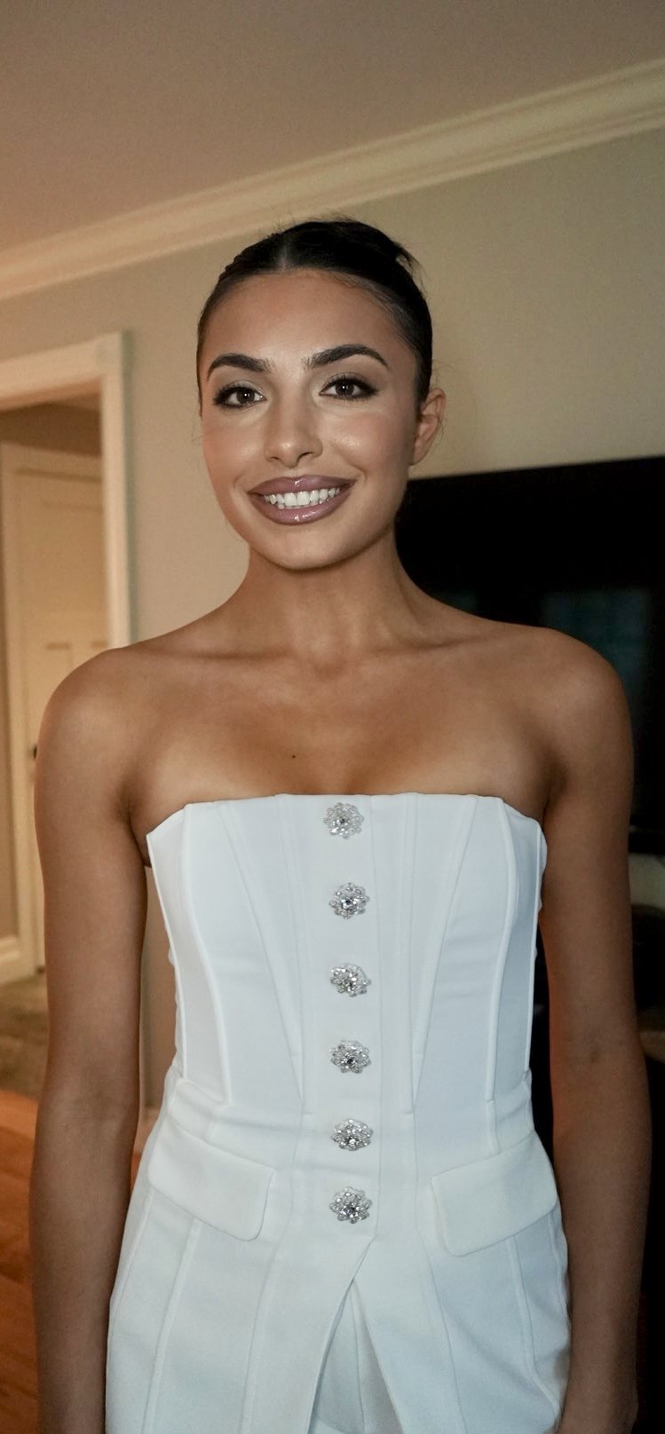 A young woman with dark hair styled in an updo, wearing a strapless white dress with decorative buttons, smiling in an indoor setting.