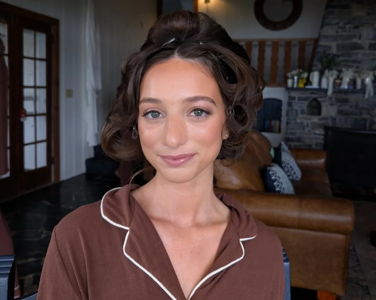 Young woman with styled brown hair and light makeup, wearing a brown shirt with white piping, in a cozy living room setting.