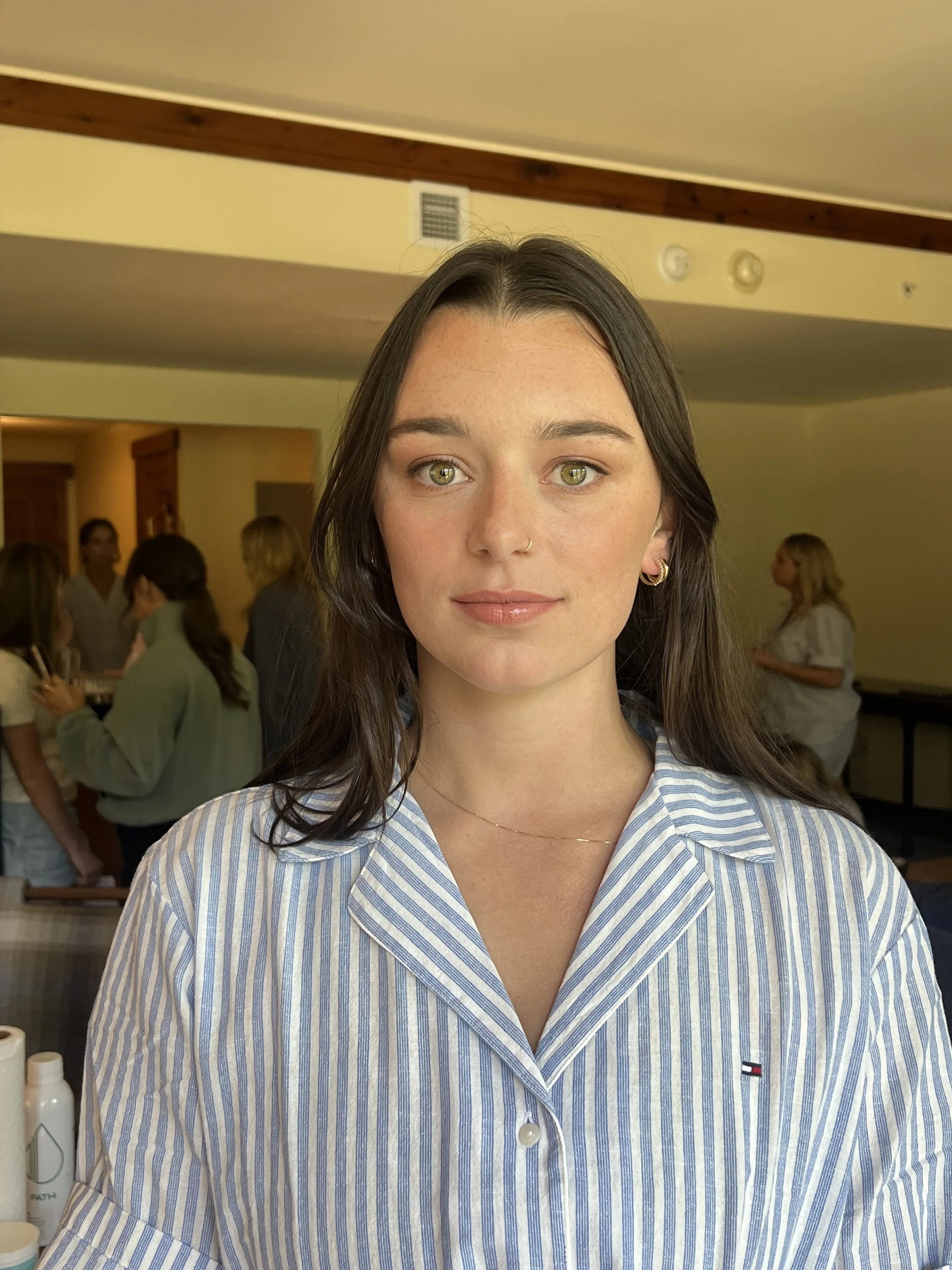 A young woman with long dark hair, green eyes, and gold jewelry, wearing a blue and white striped shirt, standing indoors with a group of people in the background.