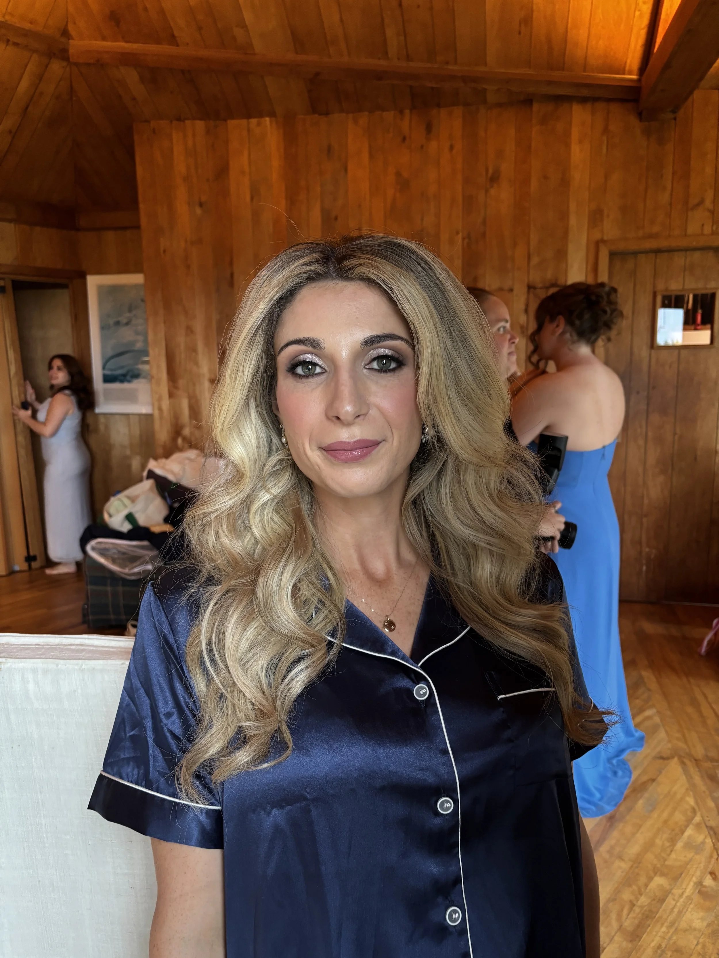 Woman with long wavy blonde hair wearing navy blue satin pajamas with white piping, sitting indoors with a wooden-paneled wall in the background.