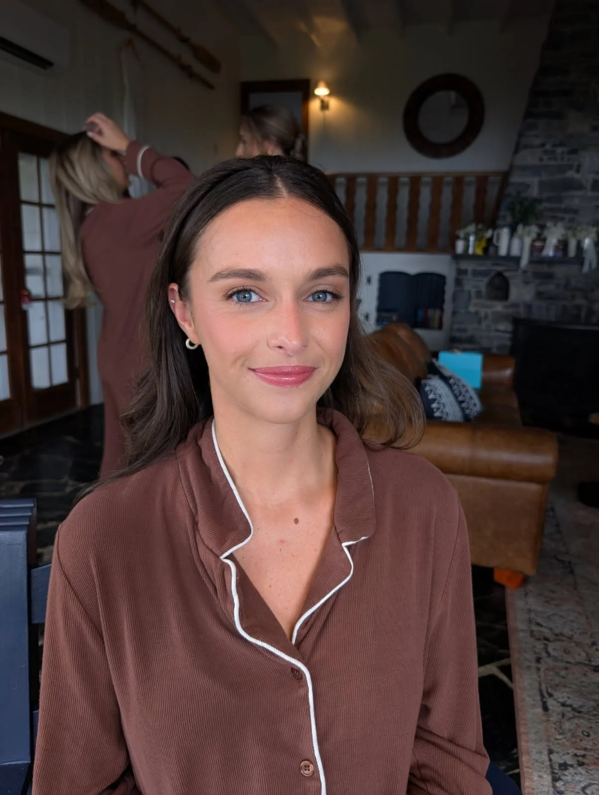 A woman with blue eyes and brown hair smiling at the camera, wearing brown pajamas with white piping, inside a cozy living room with a stone fireplace and a leather armchair.
