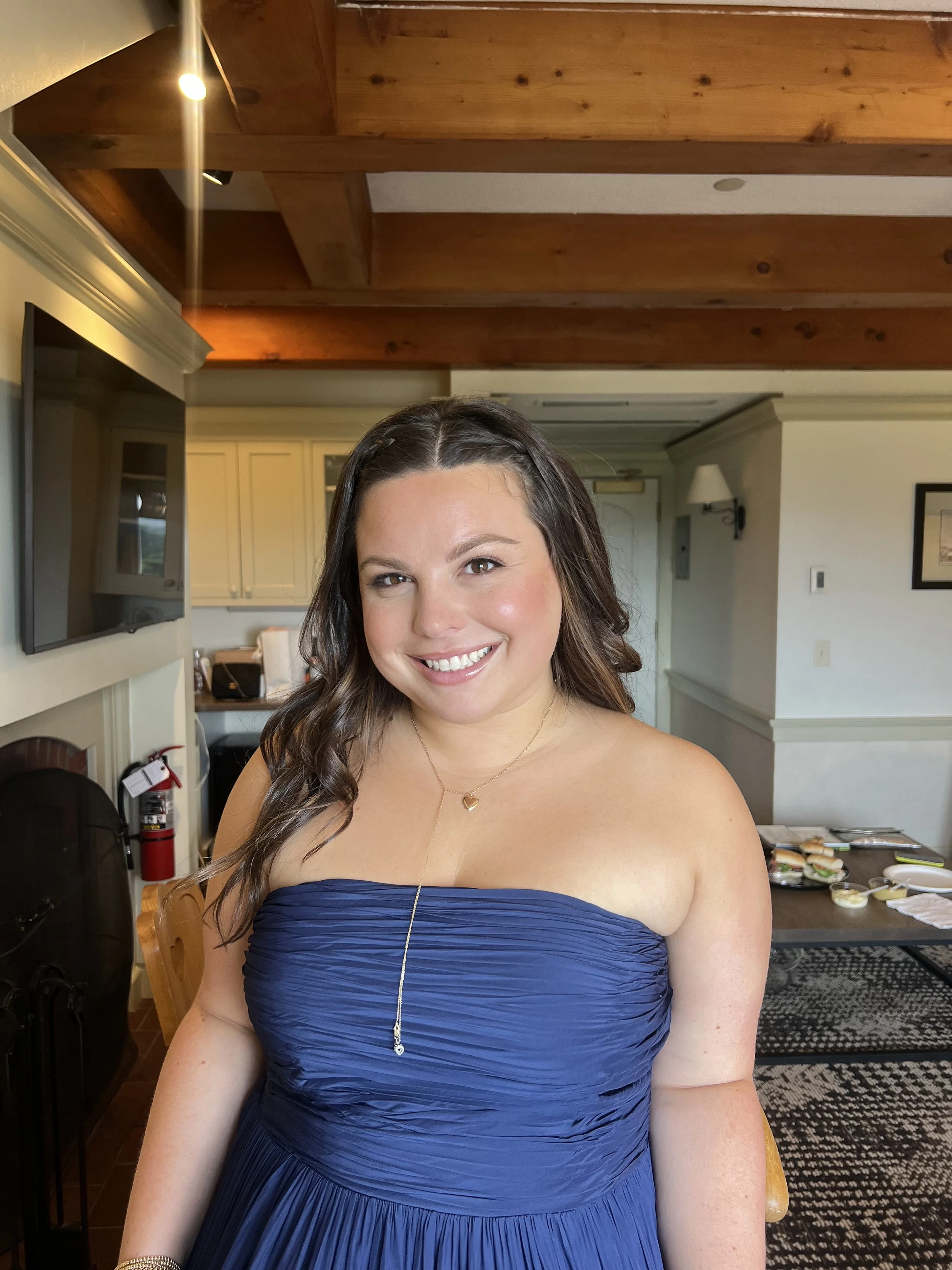 A smiling woman with long dark wavy hair wearing a strapless blue dress, standing in a cozy dining room with wooden beams and a television in the background.