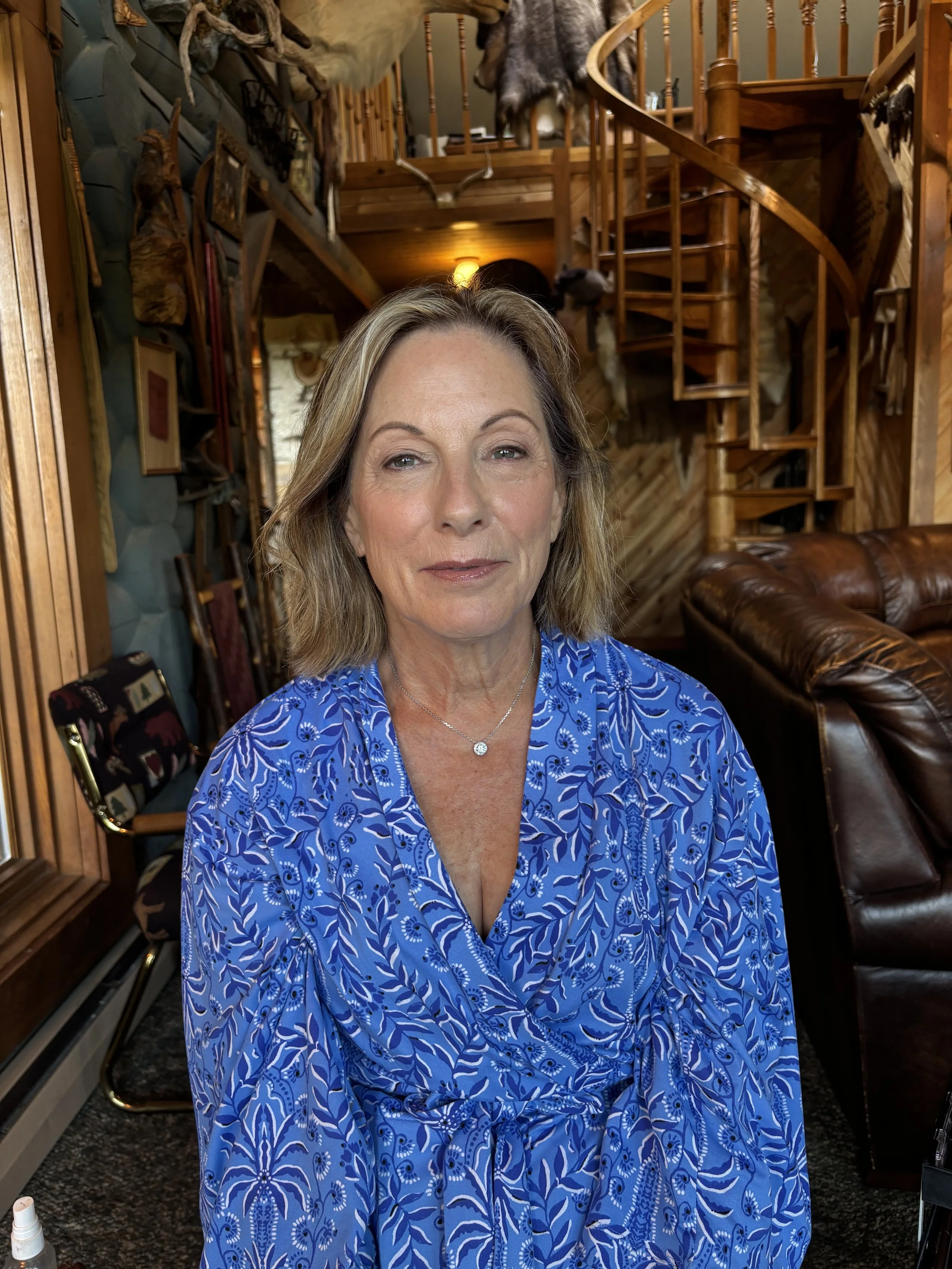 A woman with shoulder-length blonde hair and a blue patterned dress sitting in a rustic, wood-paneled room with hunting trophies and a spiral staircase in the background.