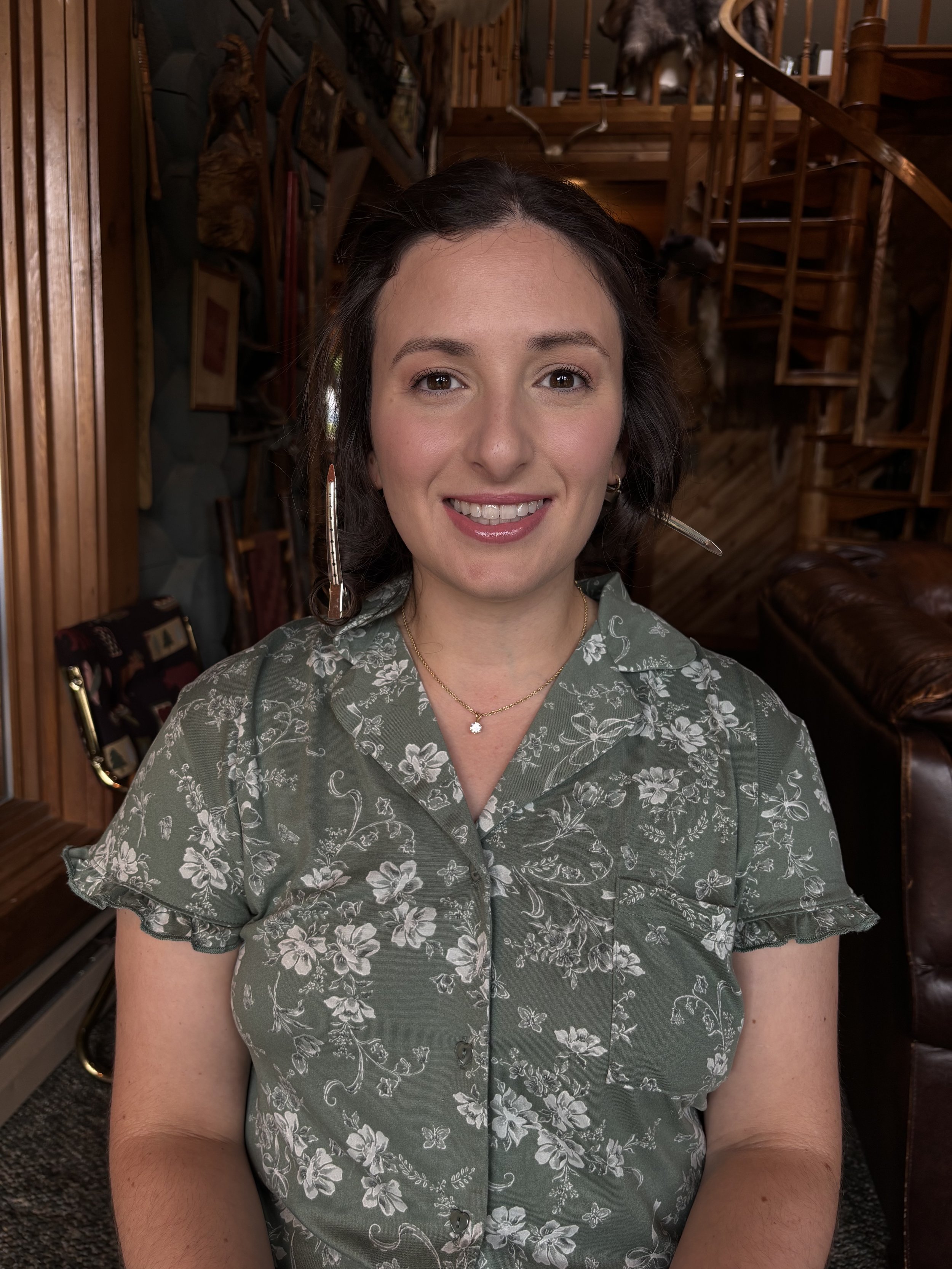 A woman with shoulder-length dark hair, smiling, wearing a green floral blouse, sitting in a wooden room with staircase and various decorations in the background.