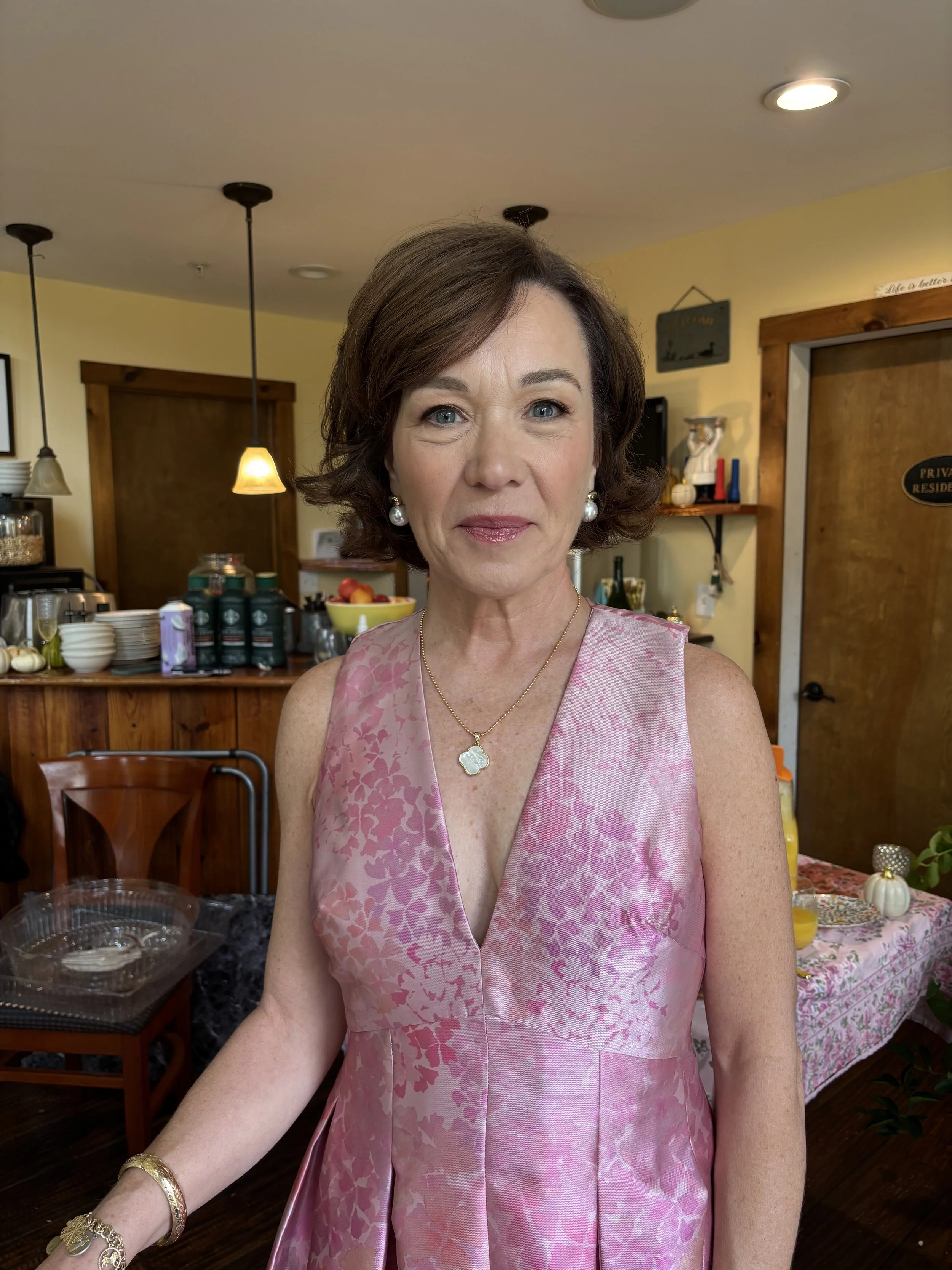 A middle-aged woman with short brown hair, wearing a pink floral sleeveless dress, pearl earrings, a gold necklace with a four-leaf clover pendant, and gold jewelry on her wrist, standing indoors in a room decorated for a celebration.