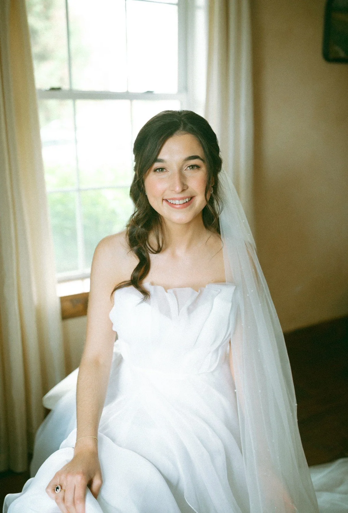 A bride in a white wedding dress sitting near a window with yellow curtains, smiling at the camera.