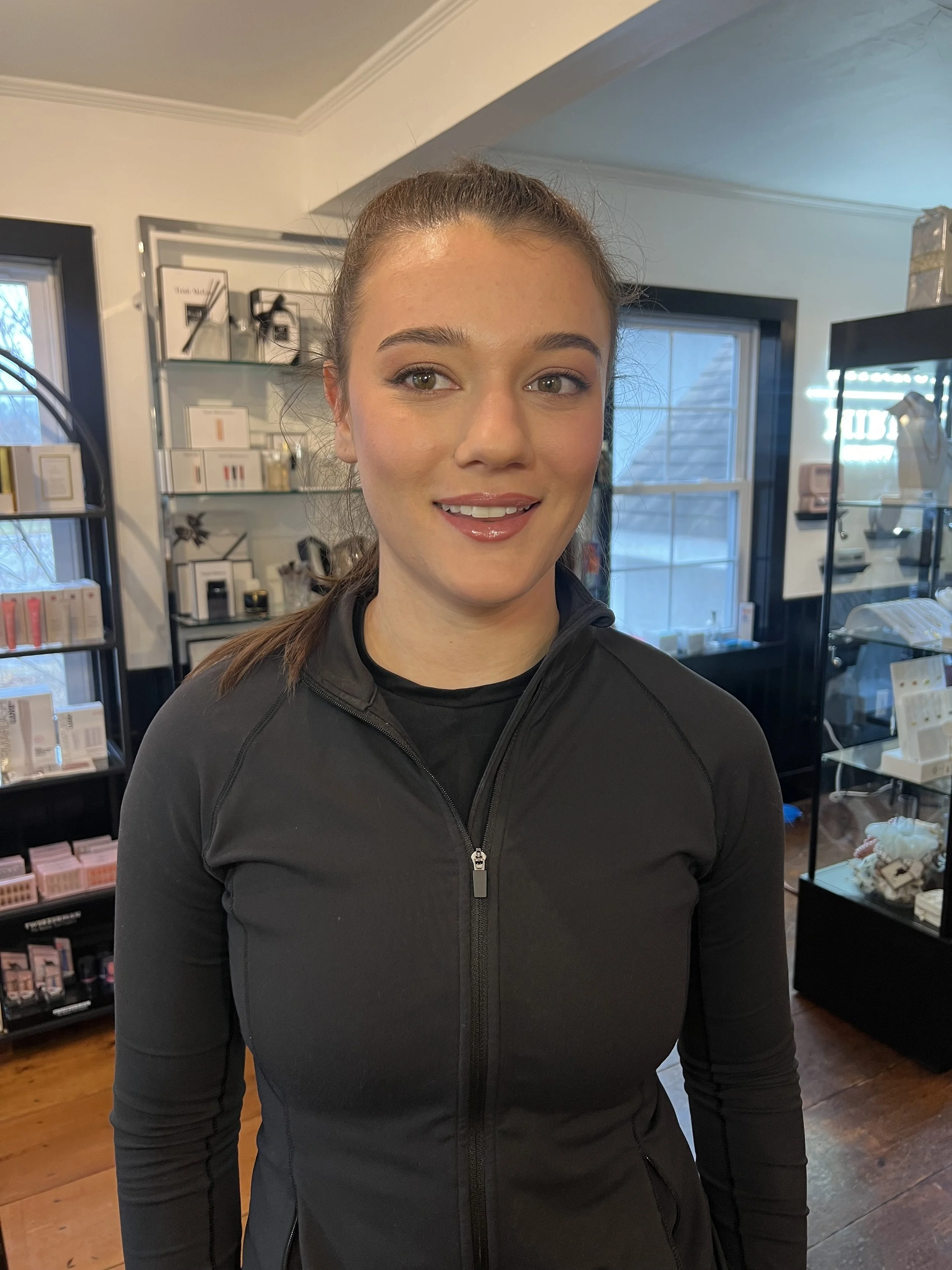A young woman with brown hair tied back, wearing a black zip-up athletic jacket, standing inside a retail or boutique store with shelves of beauty or skincare products in the background.