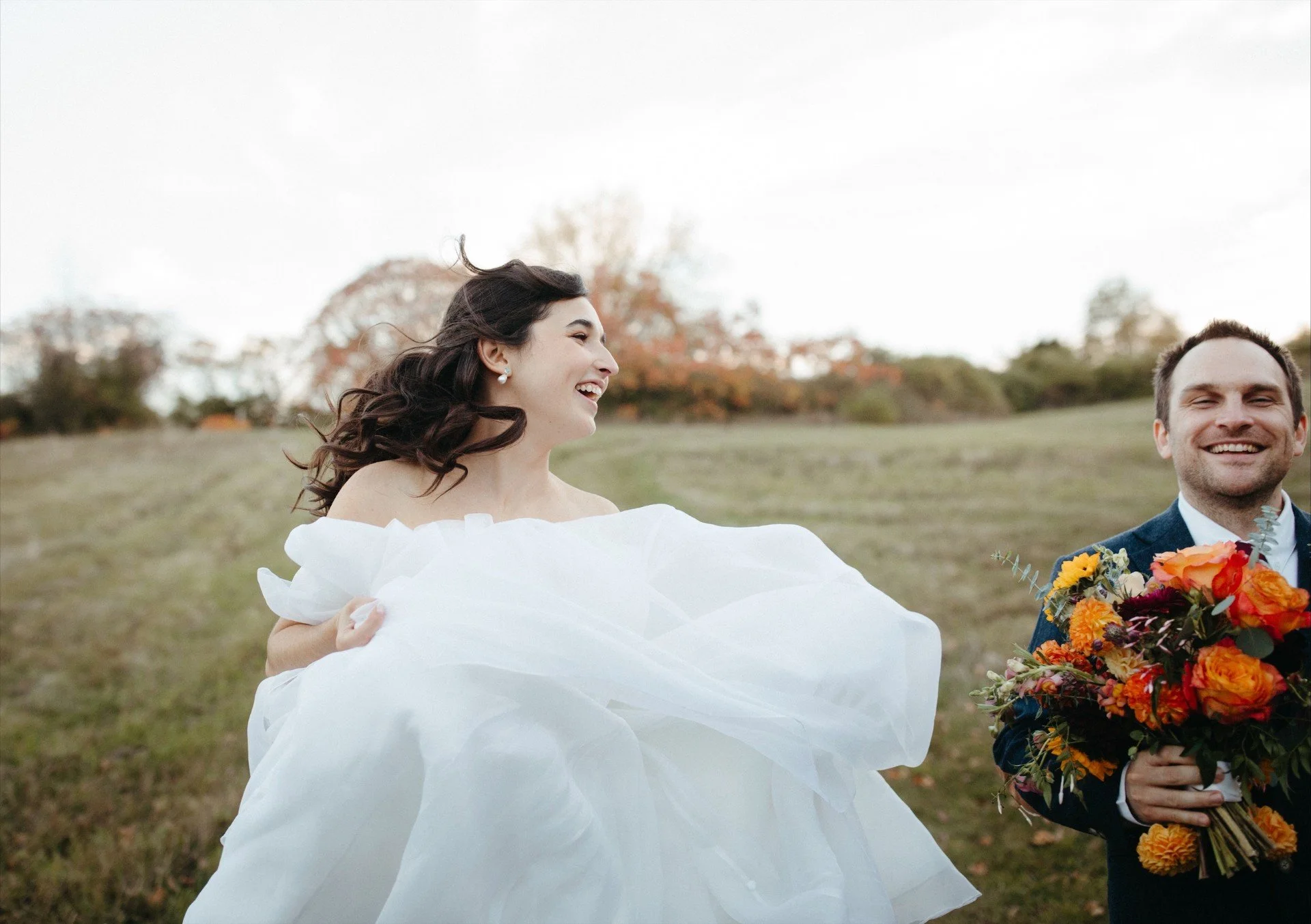A bride in a white wedding dress laughing and looking to the left, standing outdoors on grass with a groom holding a large bouquet of colorful flowers, in a park or open field with trees in fall colors.