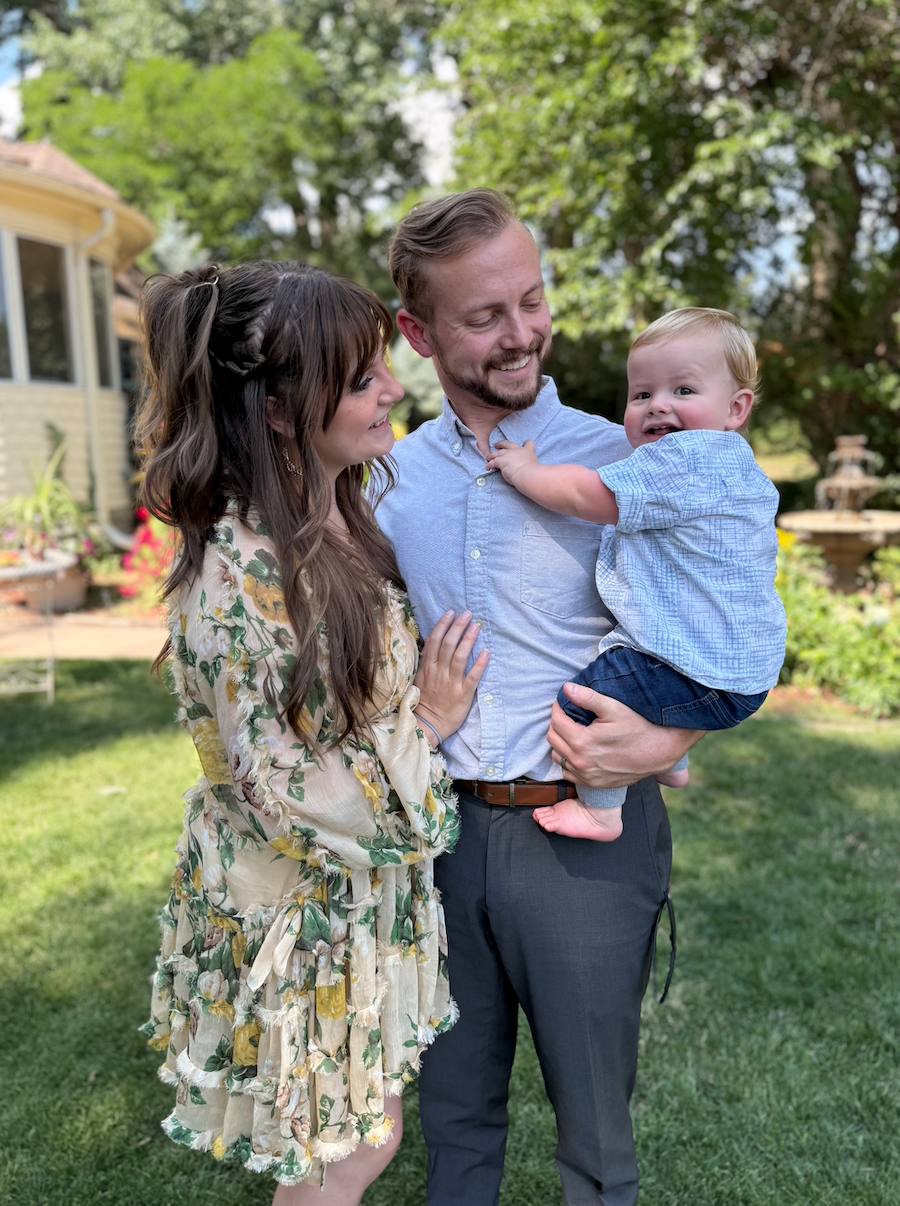 A family of three, a man holding a young child, a woman, stands outdoors in a garden with trees and a yellow house in the background. They are smiling and enjoying a sunny day.