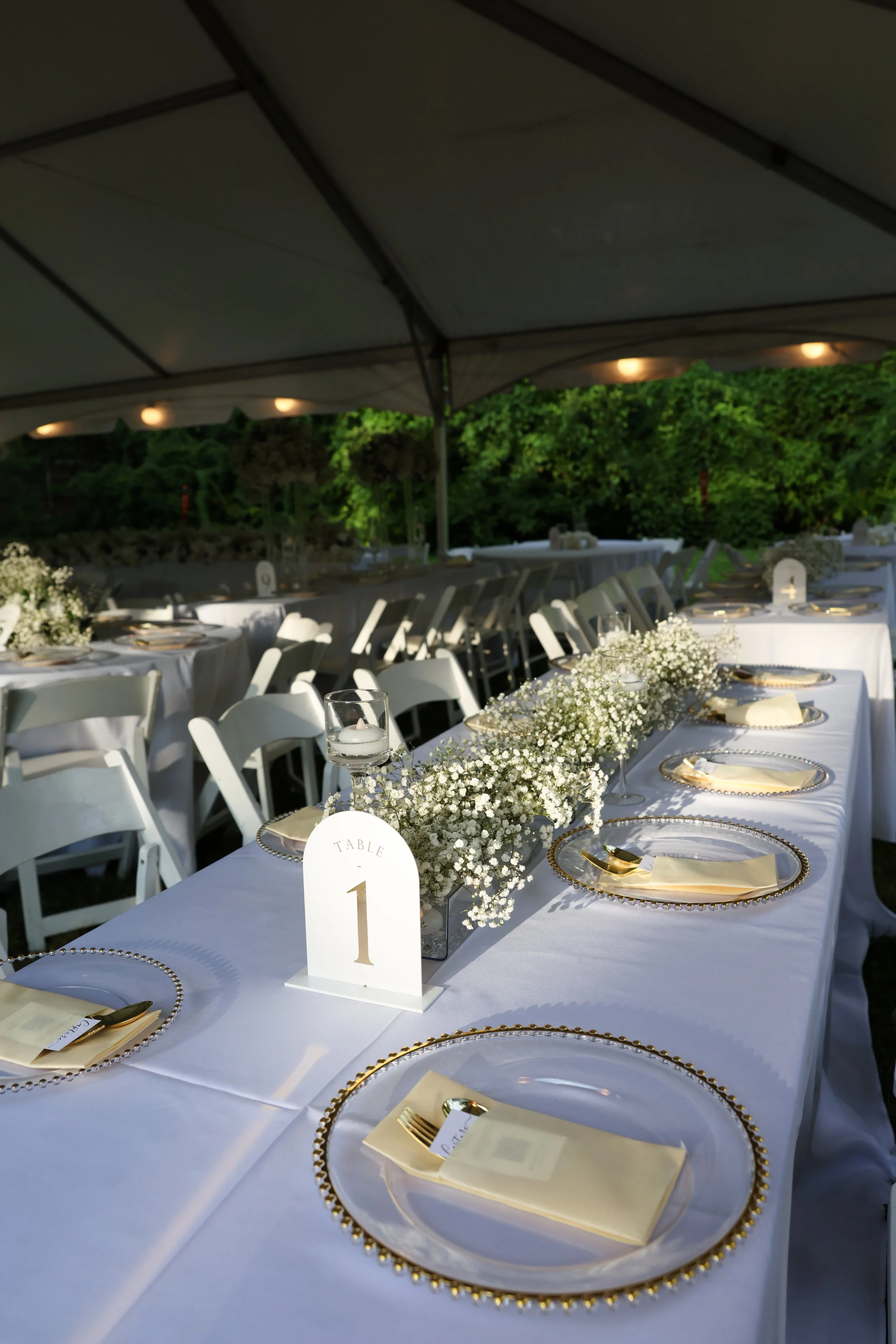 Wedding reception table set under a tent with white tablecloths, gold-rimmed plates, gold utensils, small candles, and floral centerpieces with white baby's breath.