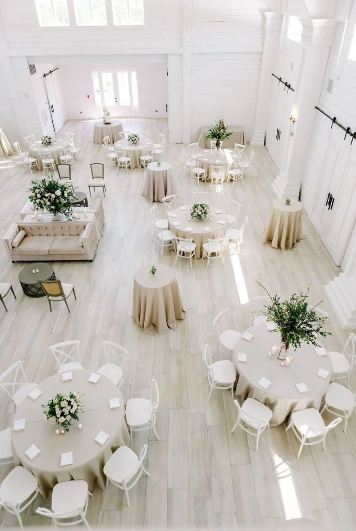Elegant white wedding reception hall with round tables covered in beige tablecloths, decorated with floral centerpieces, and white chairs. The room has white wooden walls and floors, large windows, and natural light.