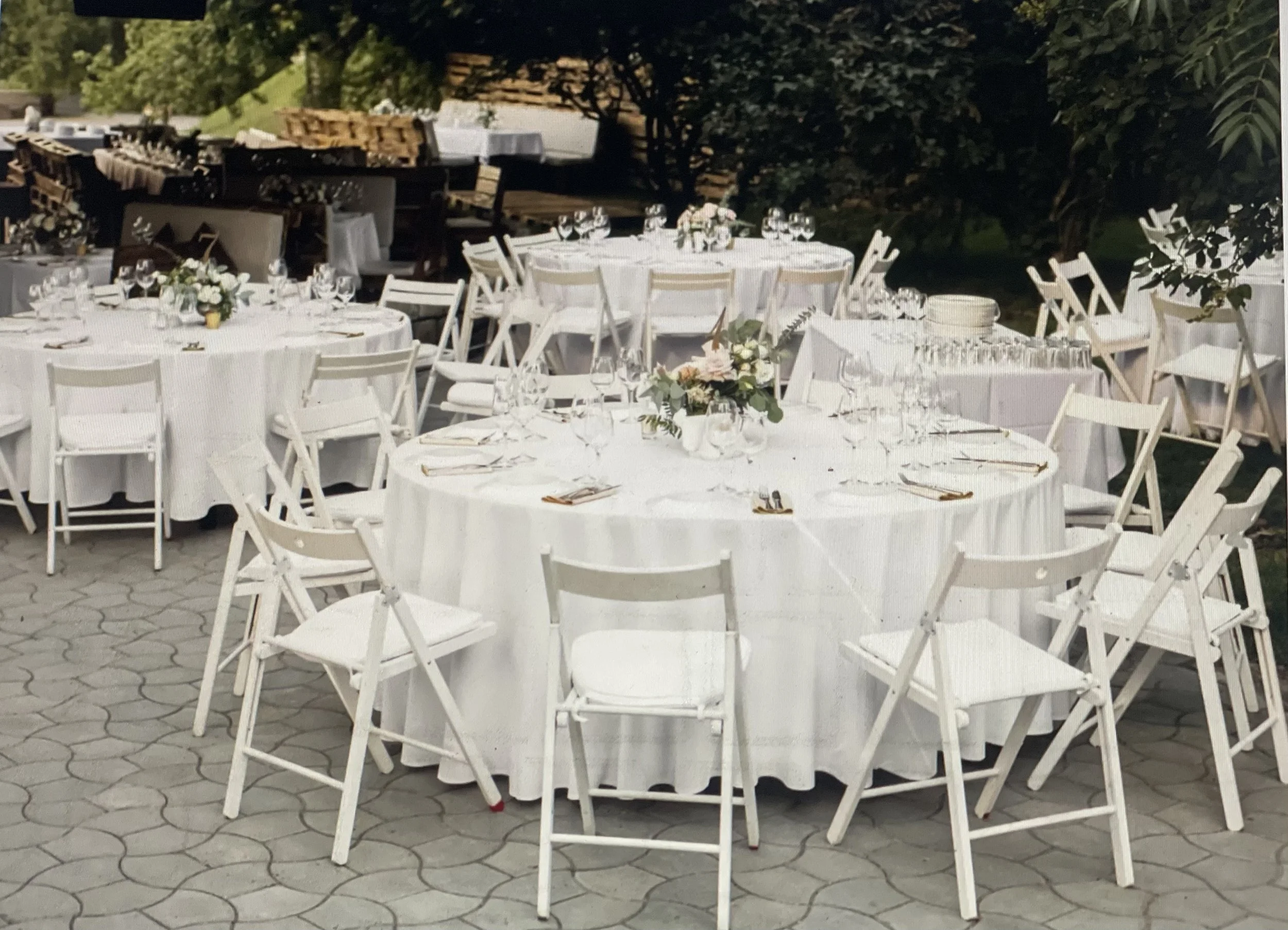 Round outdoor banquet table set with white tablecloth, wine glasses, plates, silverware, and a floral centerpiece, surrounded by white wooden folding chairs, with stacked chairs and pallets in the background.