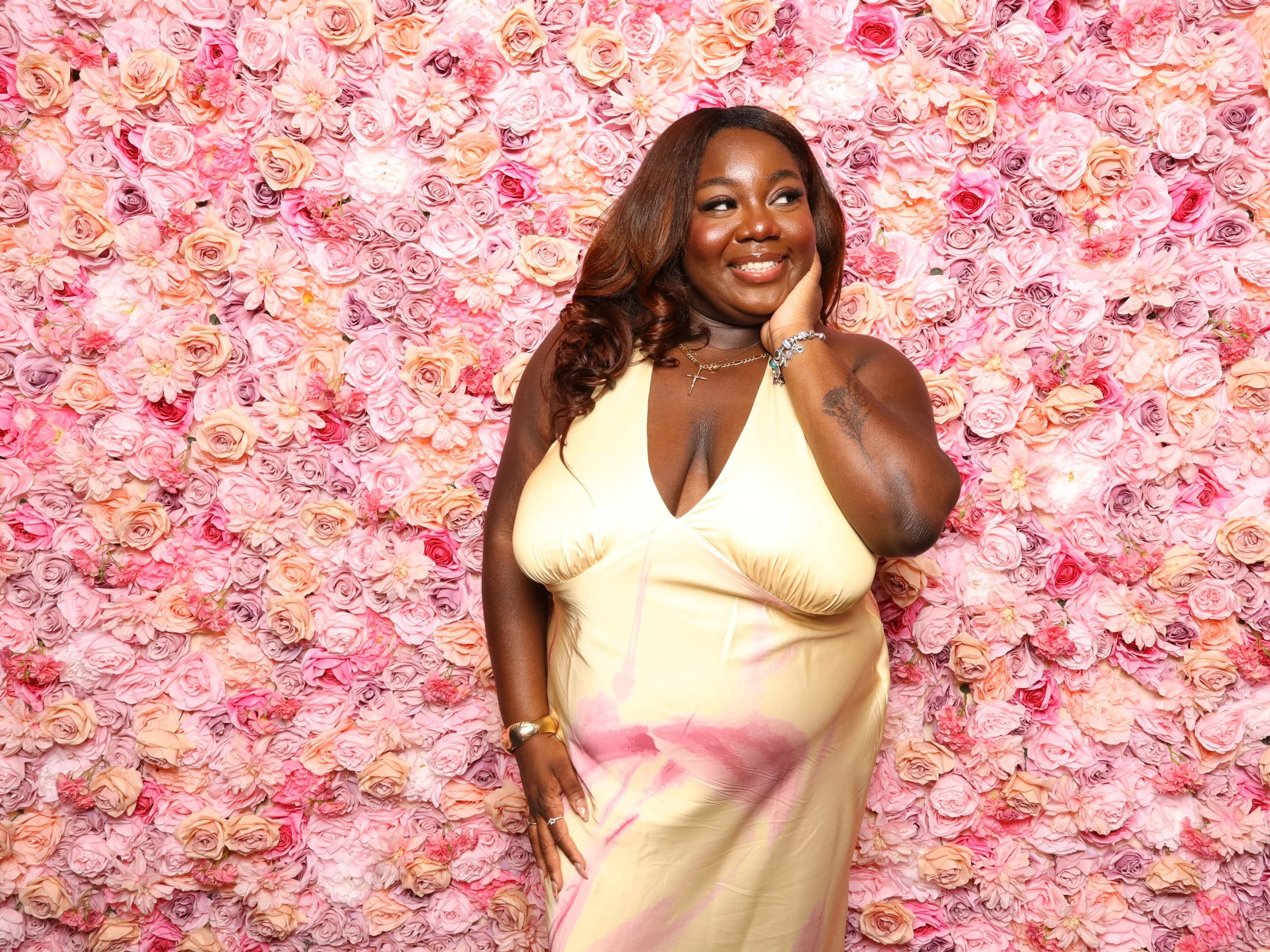 A woman in a cream-colored dress standing in front of a pink floral wall, smiling and posing with one hand resting on her cheek.