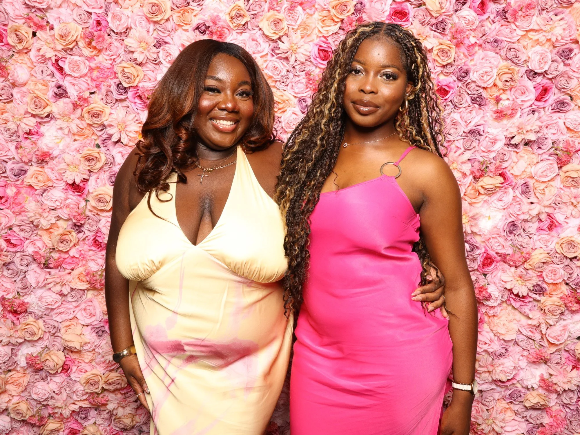 Two women smiling and posing in front of a pink floral backdrop. One woman is wearing a yellow dress, and the other is wearing a pink dress.