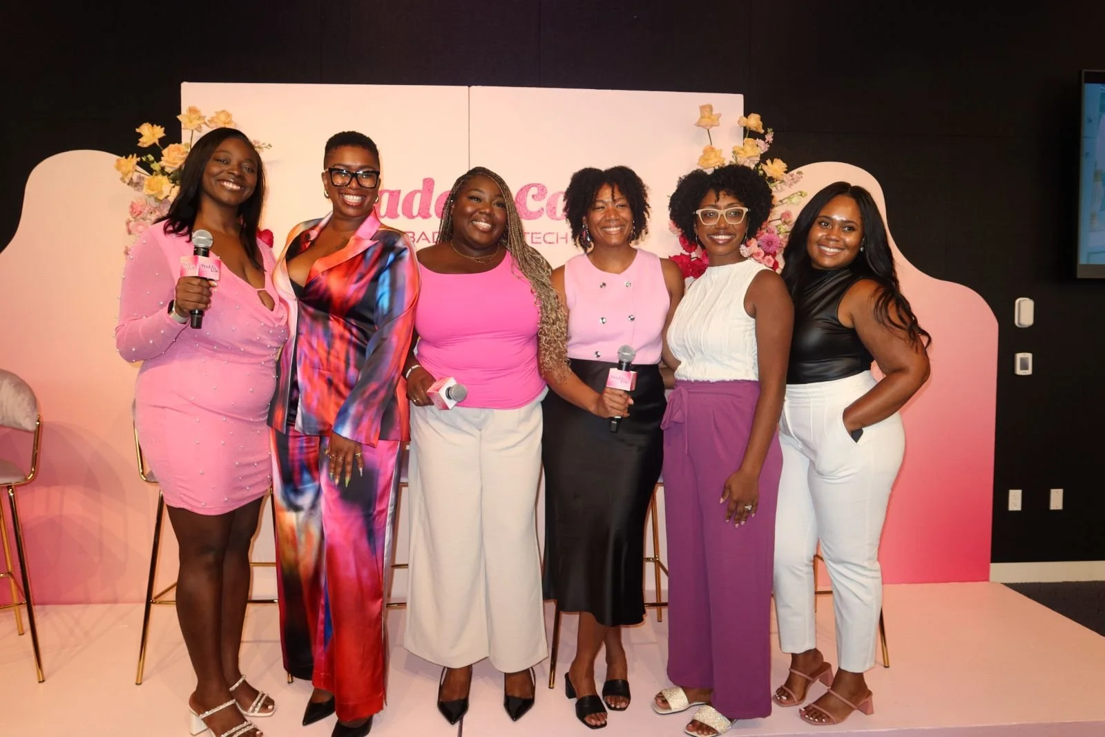 Seven women standing together on a pink stage, smiling at the camera, with some holding microphones. They are dressed in colorful stylish outfits, and behind them is a backdrop with pink and white flowers and the partial text 'GudeCo'.