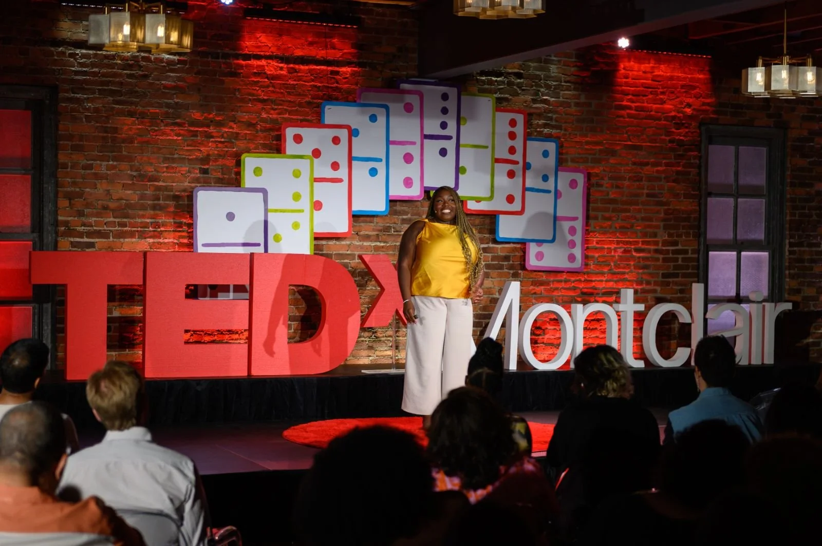 A woman standing on stage at a TEDxMontclair event, smiling with a brick wall background decorated with colorful dominoes and large TEDx and Montclair signs, and an audience seated in front.
