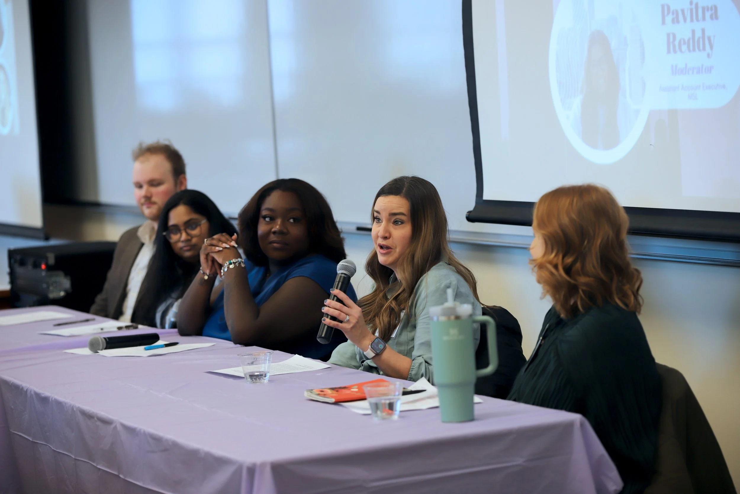 Panel of five diverse women and one man sitting at a conference table, one woman speaking into a microphone, with a presentation slide in the background.
