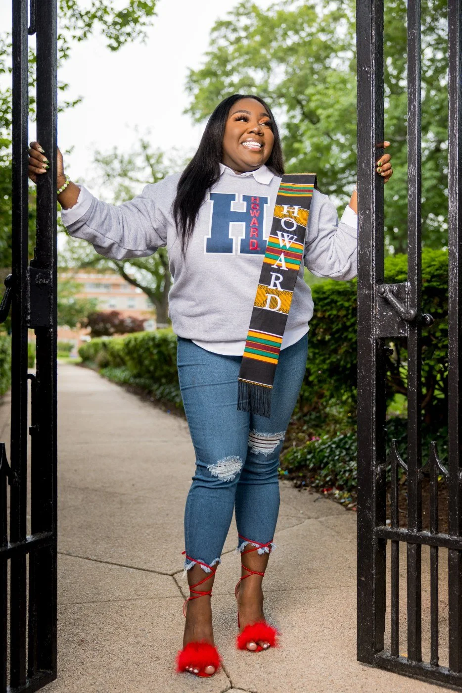 Woman standing at an open gate, smiling, wearing a graduation stole, a gray hoodie with a large letter H, and red fuzzy high heels.