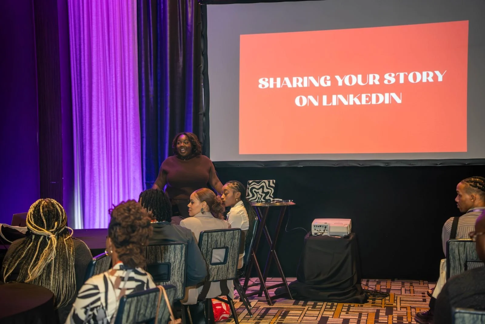A woman standing and speaking to a group of people in a conference room with purple curtains. A large screen displays the text 'Sharing Your Story on LinkedIn'.