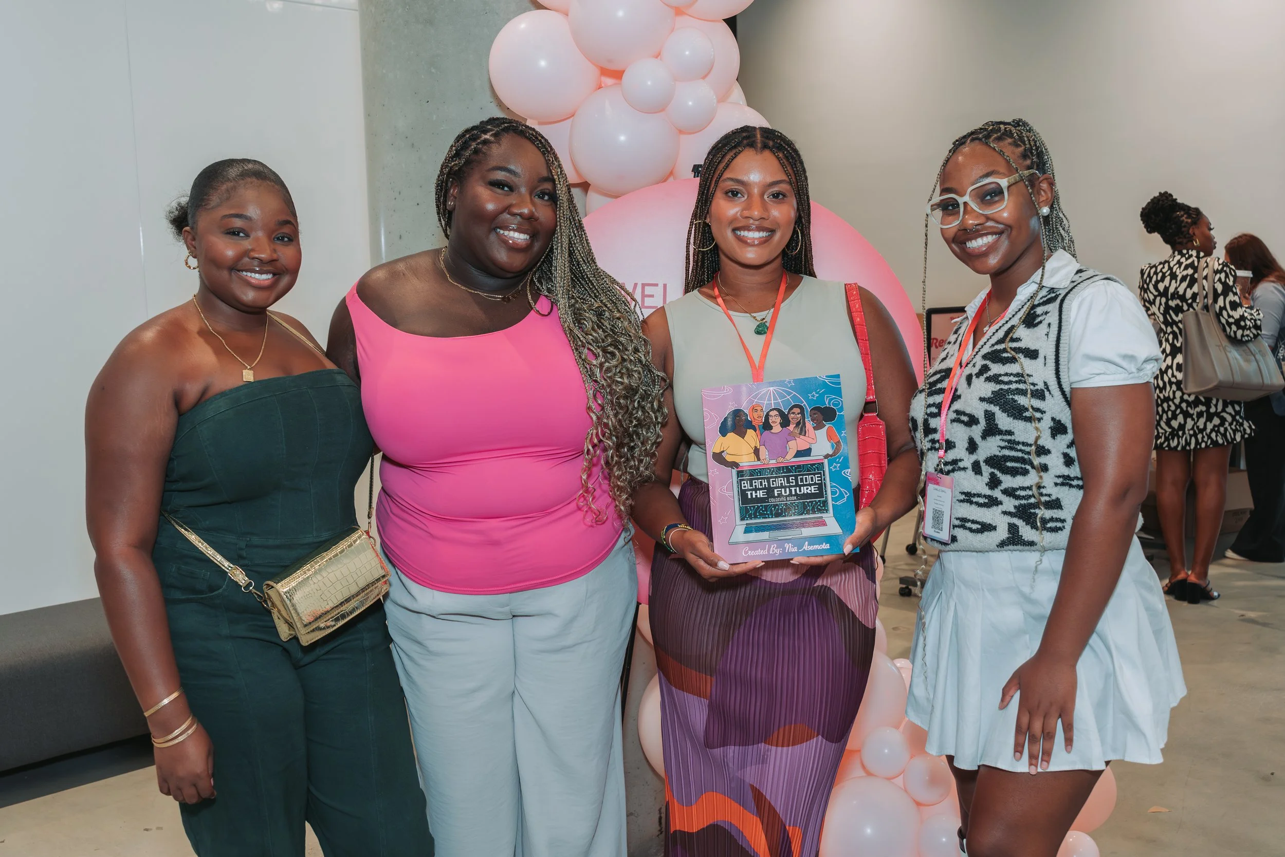 Four women standing together at an event, holding a framed sign that reads "Black Girls Code The Future," with pink balloons in the background.
