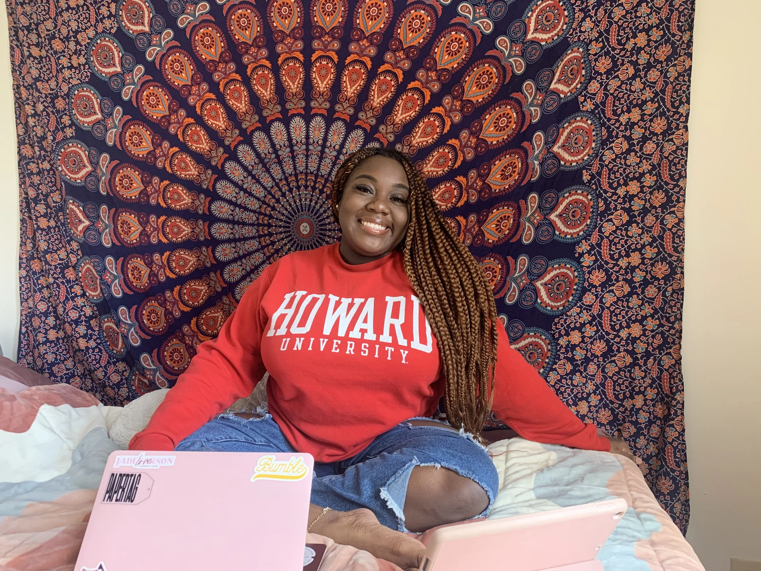 A smiling woman with long braided hair sitting on a bed in front of a decorative tapestry with orange and blue patterns, wearing a red Howard University sweatshirt and ripped jeans.