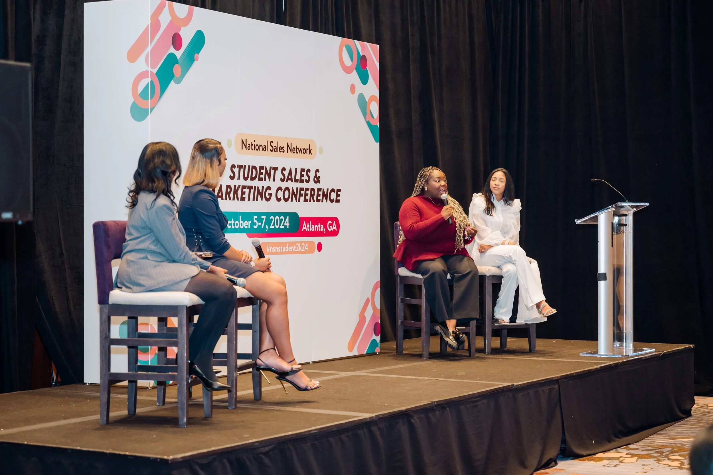 Four women sitting on a stage at a conference, with a large event banner behind them, discussing during the Student Sales & Marketing Conference in Atlanta, GA, October 5-7, 2024.