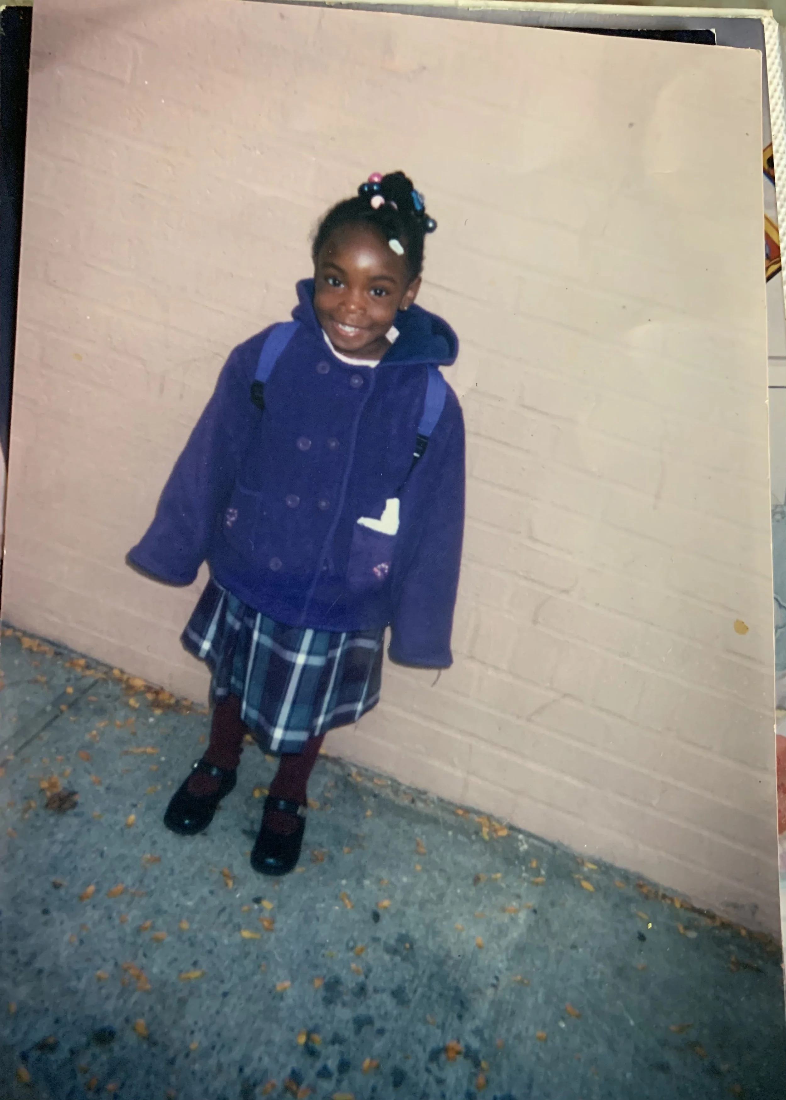 A young girl with braided hair decorated with colorful beads, wearing a blue coat, plaid skirt, maroon tights, and black shoes, stands outside against a beige brick wall, smiling at the camera.