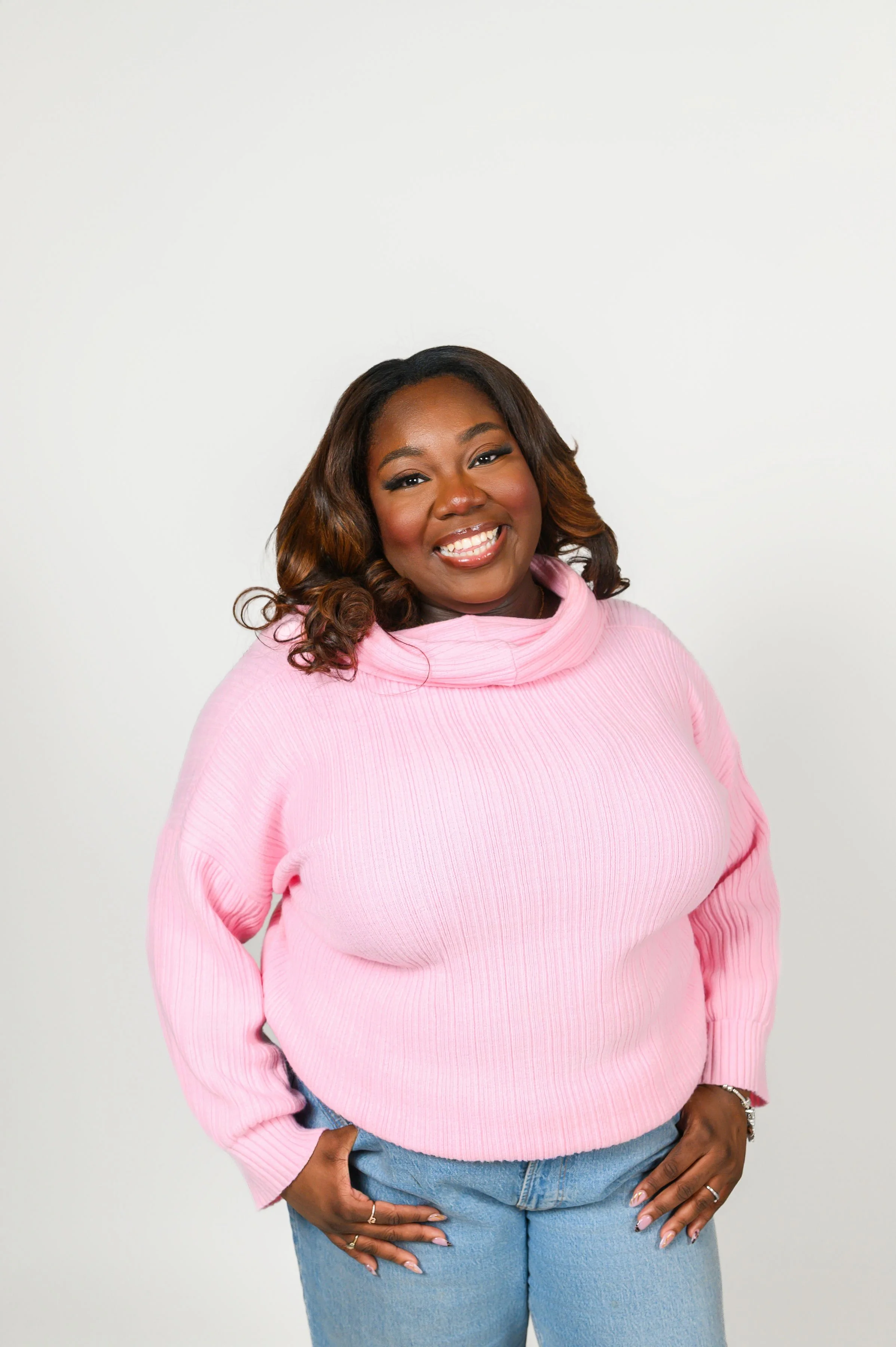 A woman with medium-dark skin tone, curly brown hair with highlights, smiling, wearing a pink sweater and light blue jeans, standing against a plain white background.