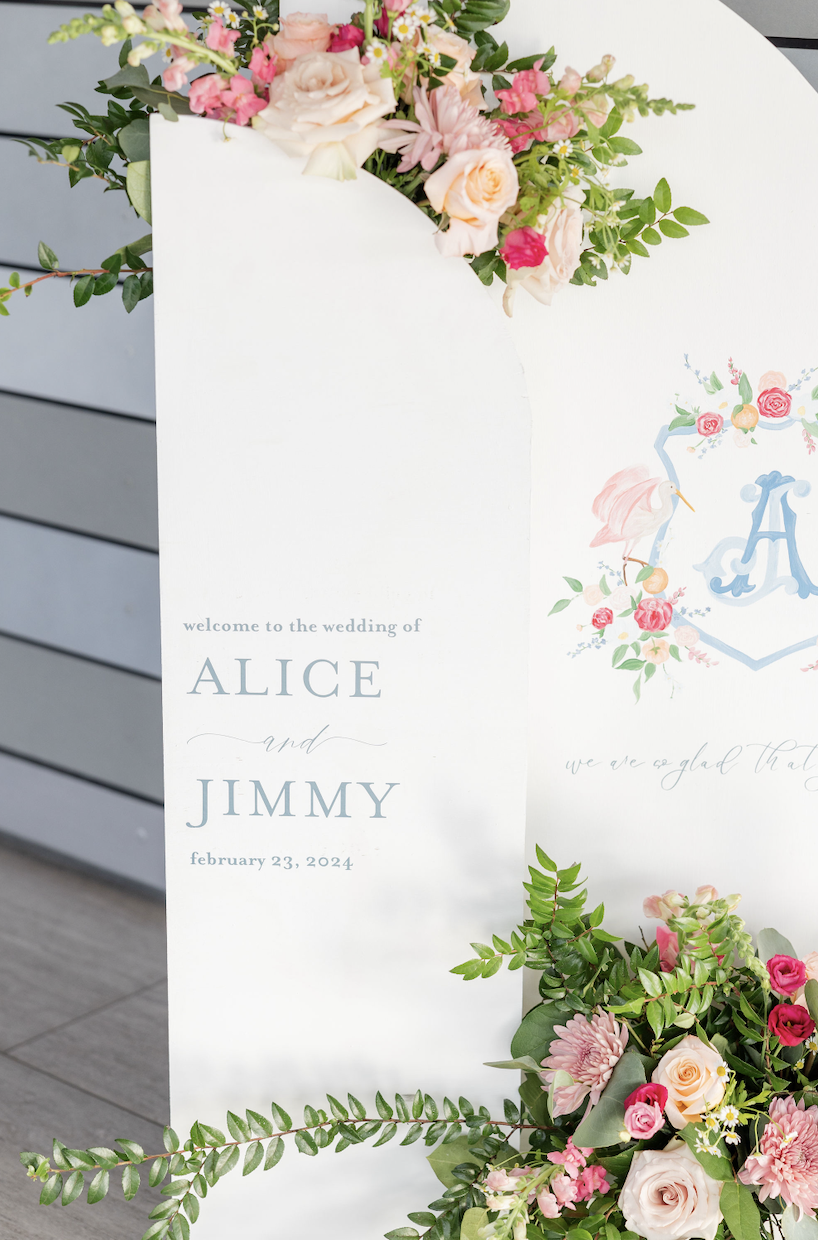 Wedding welcome sign with floral arrangements on top and bottom, featuring pink, peach, and white roses, greenery, and other flowers. The sign reads "welcome to the wedding of Alice and Jimmy February 23, 2024".