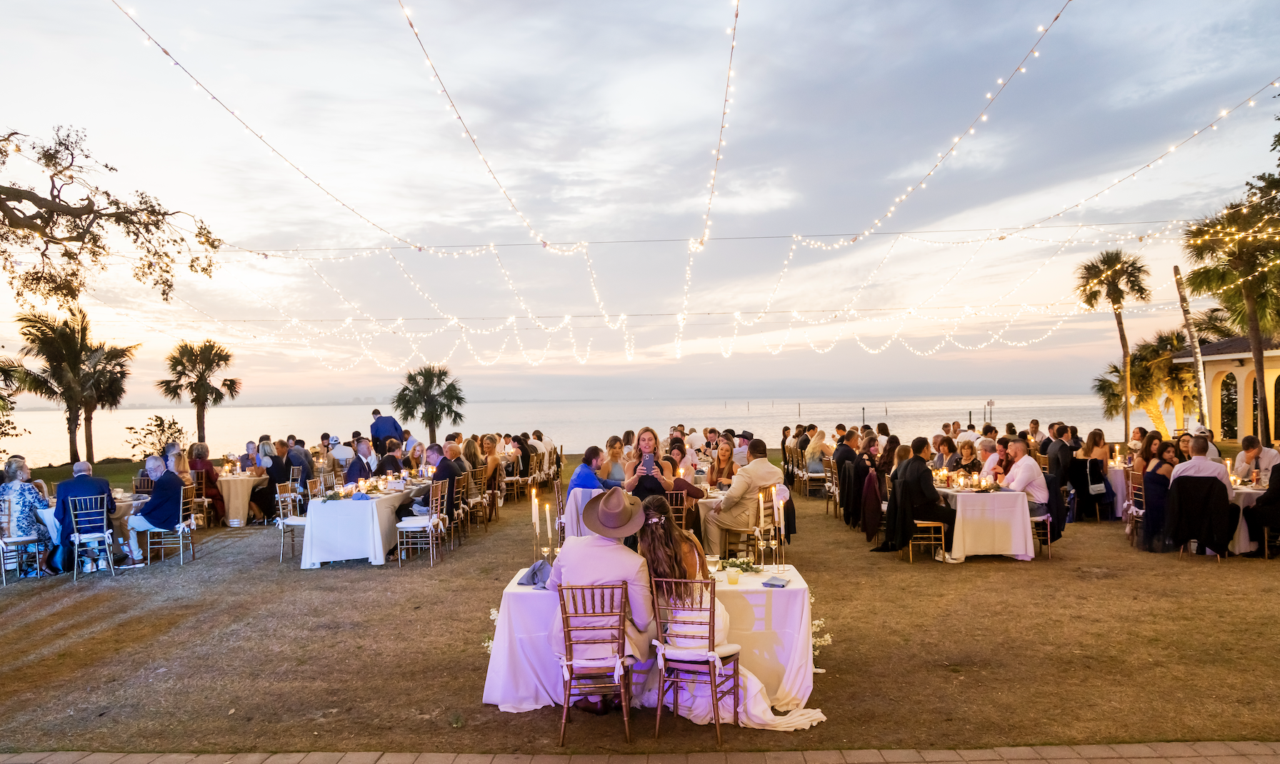 Outdoor wedding reception at sunset with string lights, palm trees, and a view of the water, featuring guests seated at tables, and a couple seated at the head table facing the water.