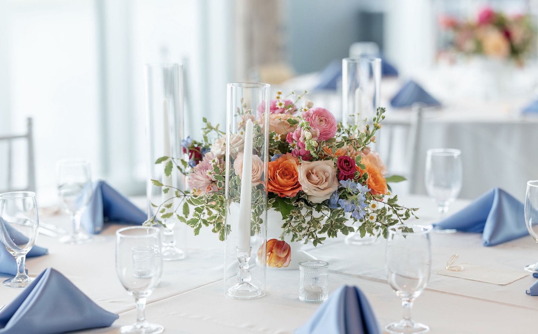 A table set for a formal event with a floral centerpiece featuring pink, orange, cream, and purple flowers, surrounded by glassware and blue cloth napkins.